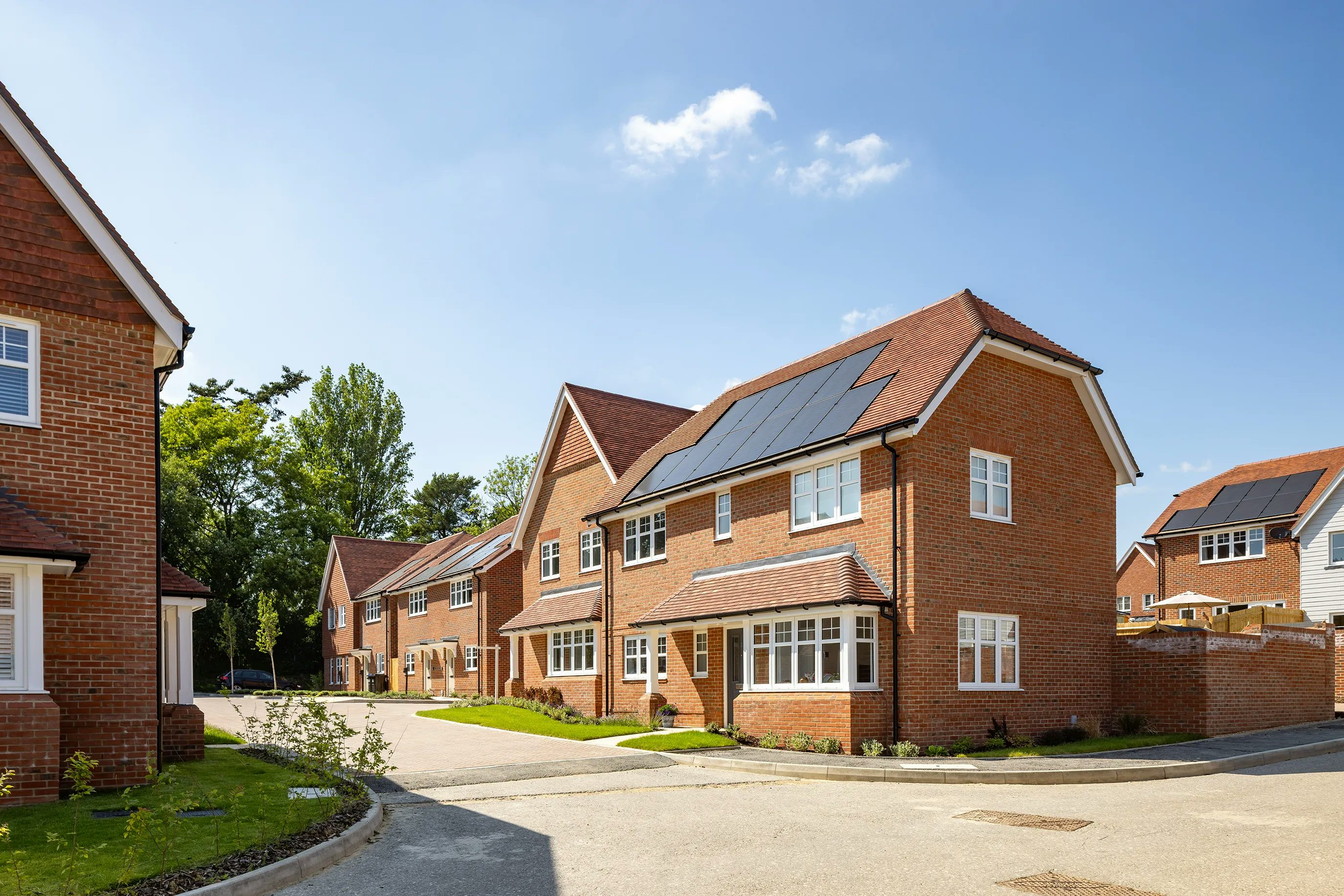 A sunny suburban street lined with modern two-story brick houses, some featuring solar panels on their pitched roofs. Neatly trimmed lawns border a paved road, while a backdrop of green trees and a few fluffy clouds enhances the sense of a peaceful, eco-conscious neighborhood.