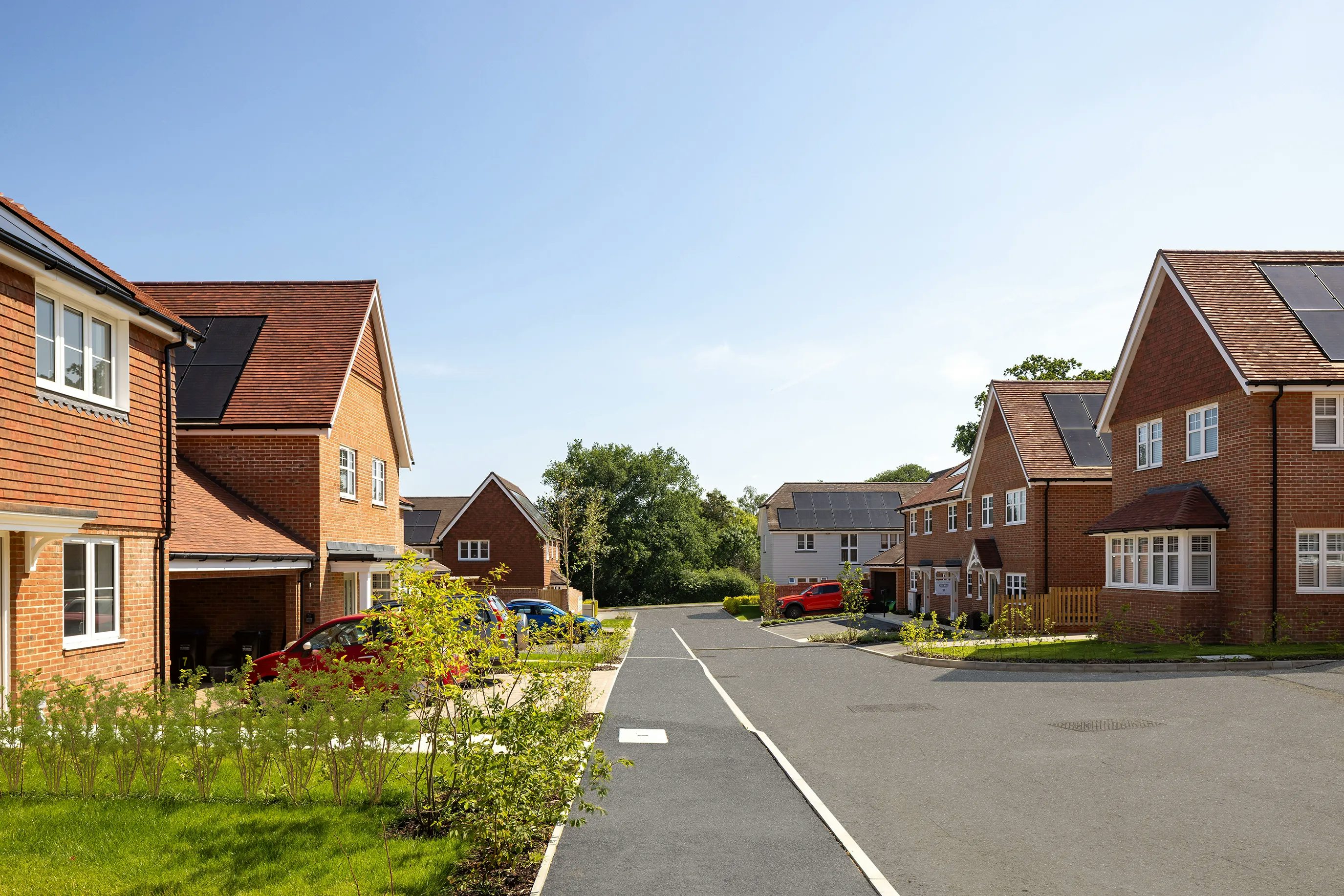 A clean, sunlit suburban street flanked by modern detached brick houses, some topped with solar panels. Driveways feature parked cars, and neatly trimmed lawns and trees line the pavement. The clear blue sky adds to the sense of order and calm in this environmentally conscious neighbourhood.