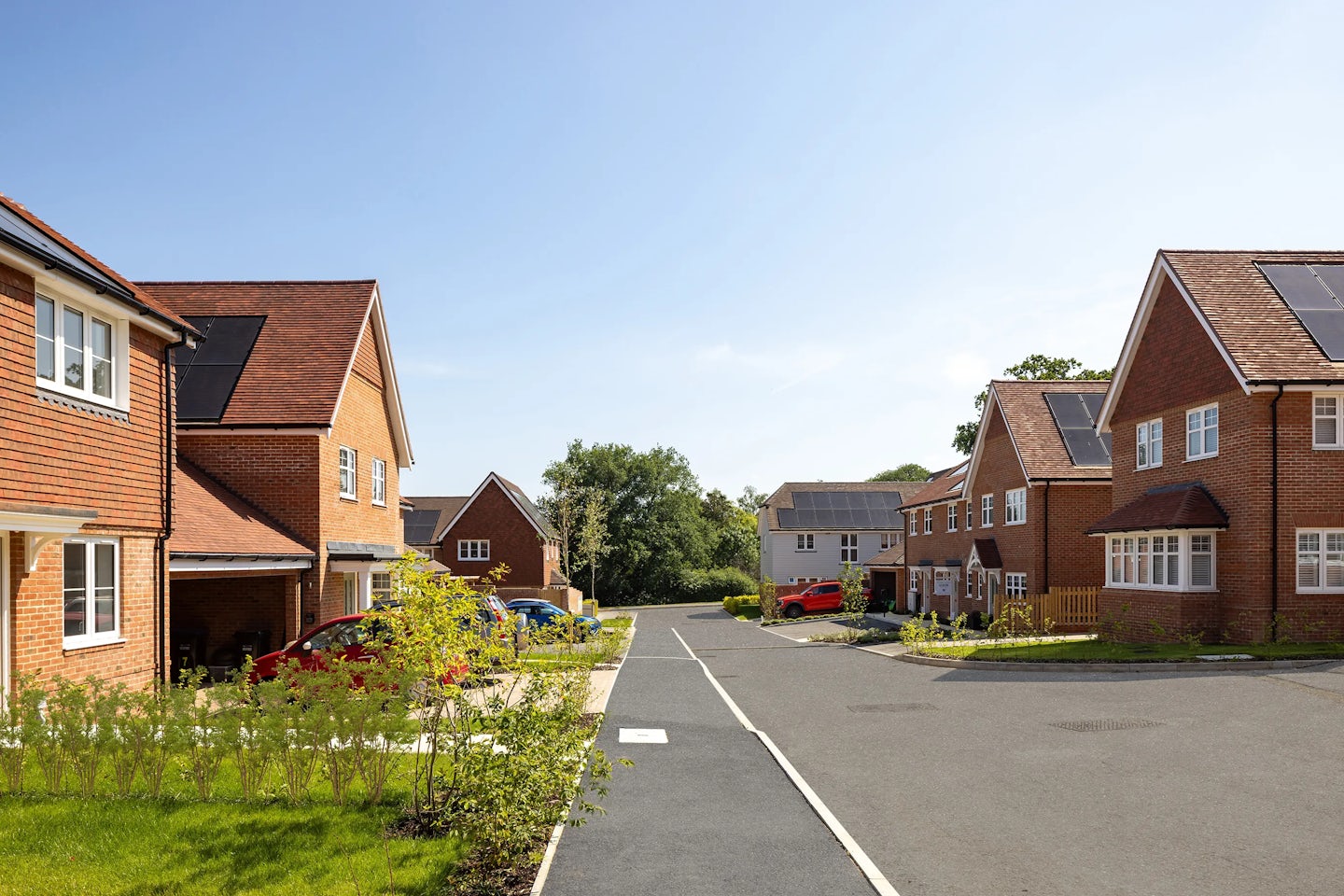 A clean, sunlit suburban street flanked by modern detached brick houses, some topped with solar panels. Driveways feature parked cars, and neatly trimmed lawns and trees line the pavement. The clear blue sky adds to the sense of order and calm in this environmentally conscious neighbourhood.