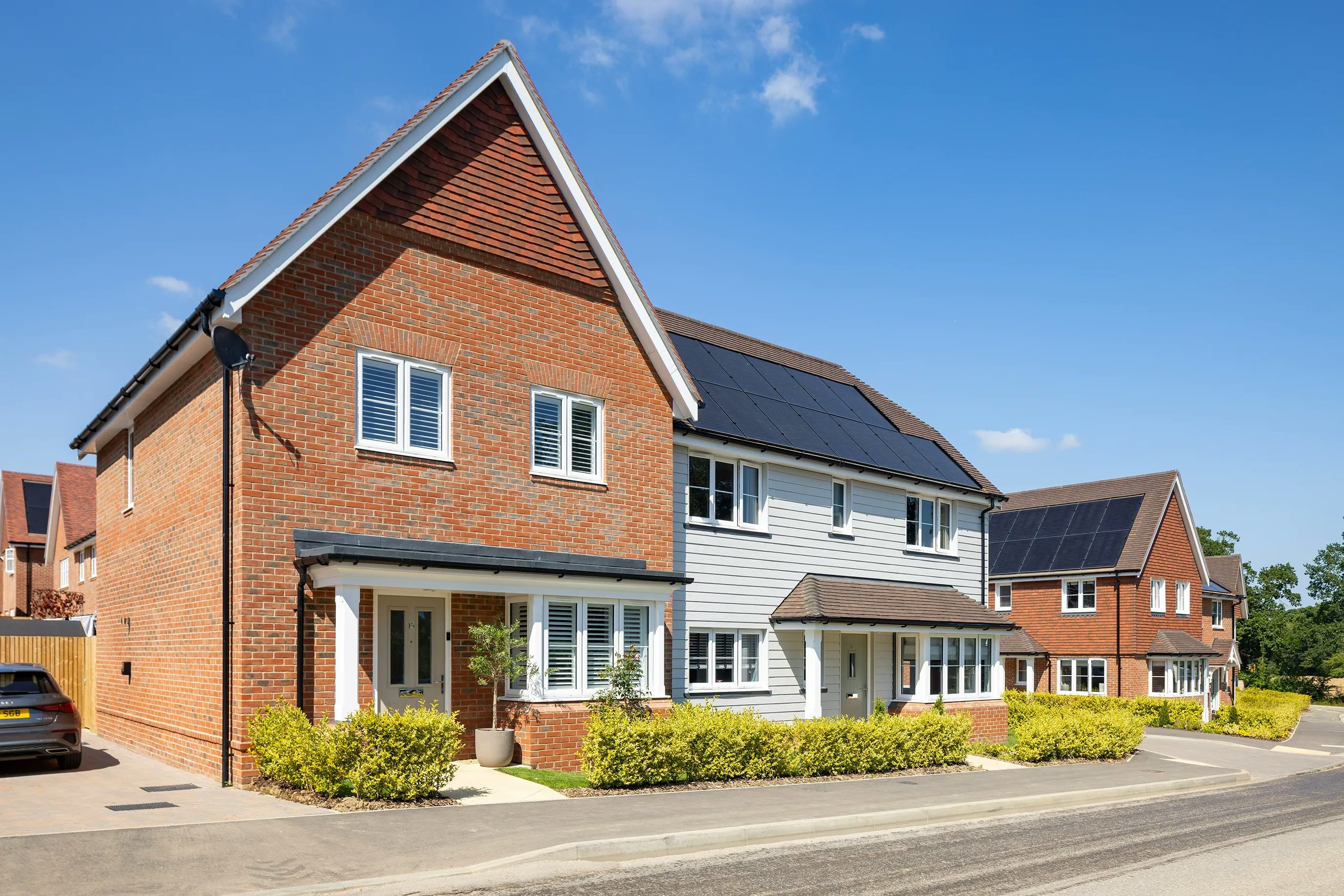 A row of contemporary suburban houses featuring a mix of white siding and warm-toned brick façades, each with large windows and tidy front gardens. Rooftops are fitted with solar panels, reflecting a focus on sustainability. The setting is calm and sunlit beneath a partly cloudy blue sky.