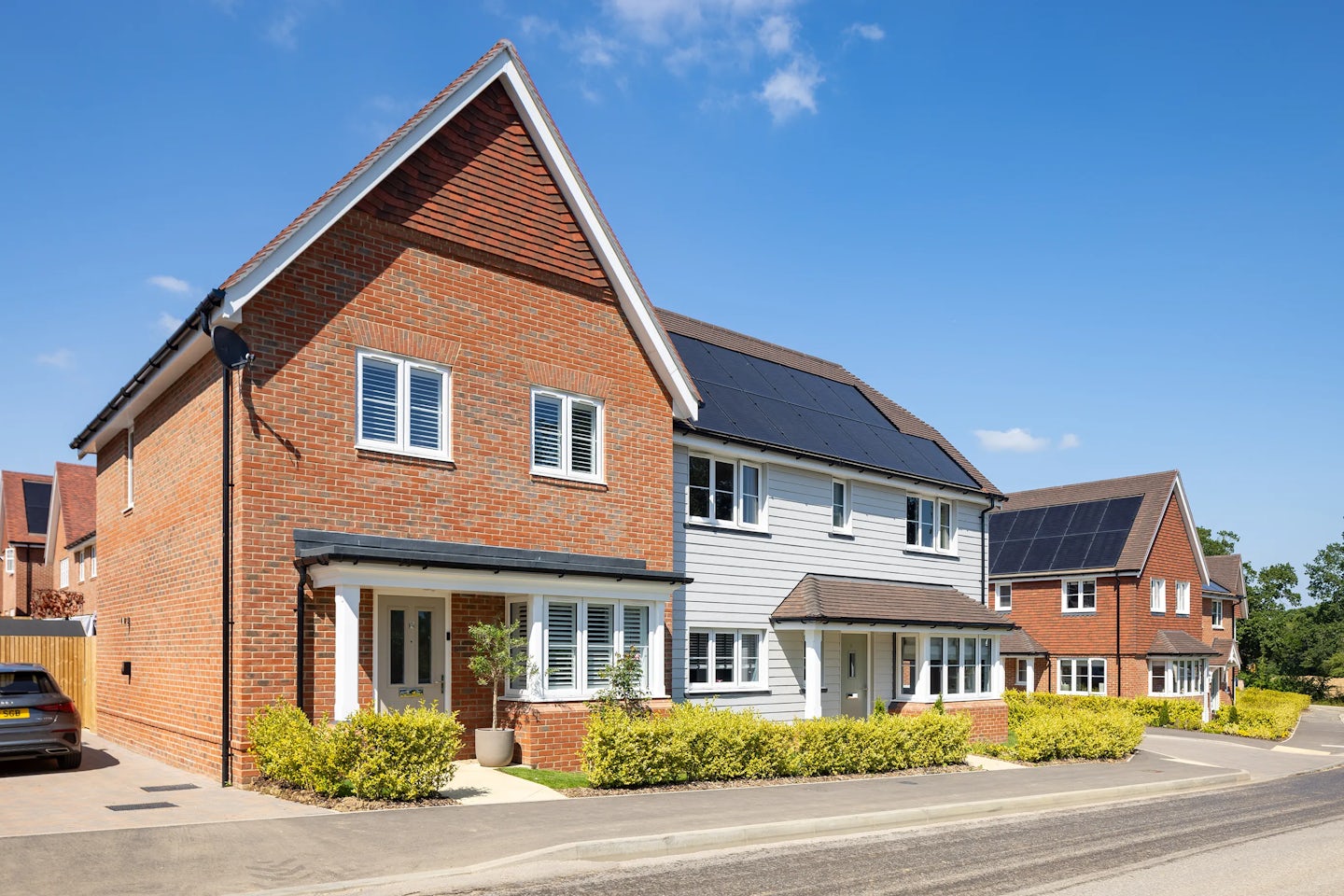 A row of contemporary suburban houses featuring a mix of white siding and warm-toned brick façades, each with large windows and tidy front gardens. Rooftops are fitted with solar panels, reflecting a focus on sustainability. The setting is calm and sunlit beneath a partly cloudy blue sky.