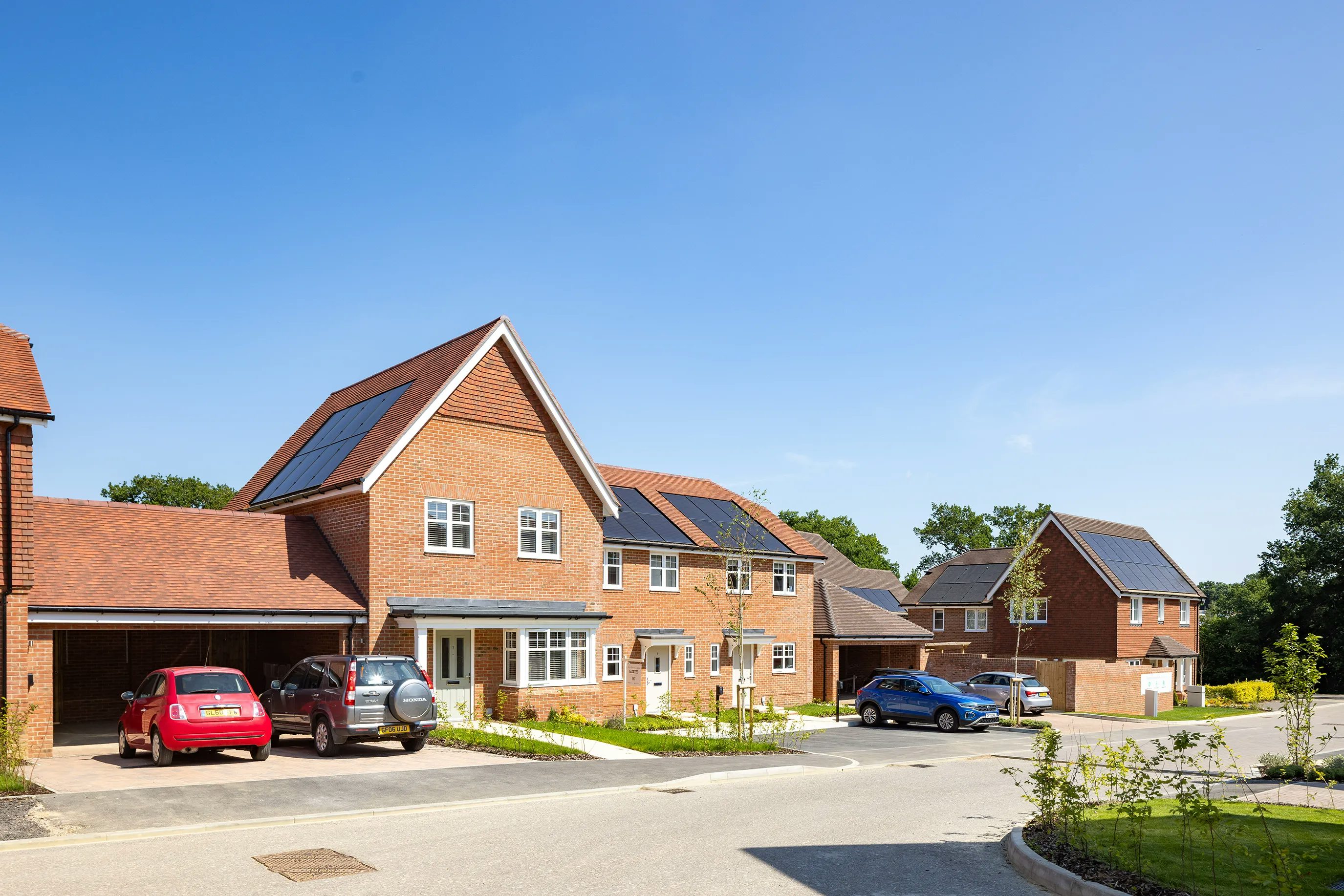 A sunny suburban street lined with modern brick houses, each featuring pitched roofs with solar panels. Cars are parked in driveways and along the road. Neatly trimmed lawns and young trees enhance the clean, well-maintained appearance of the neighborhood. The clear blue sky emphasizes a bright, environmentally conscious atmosphere.