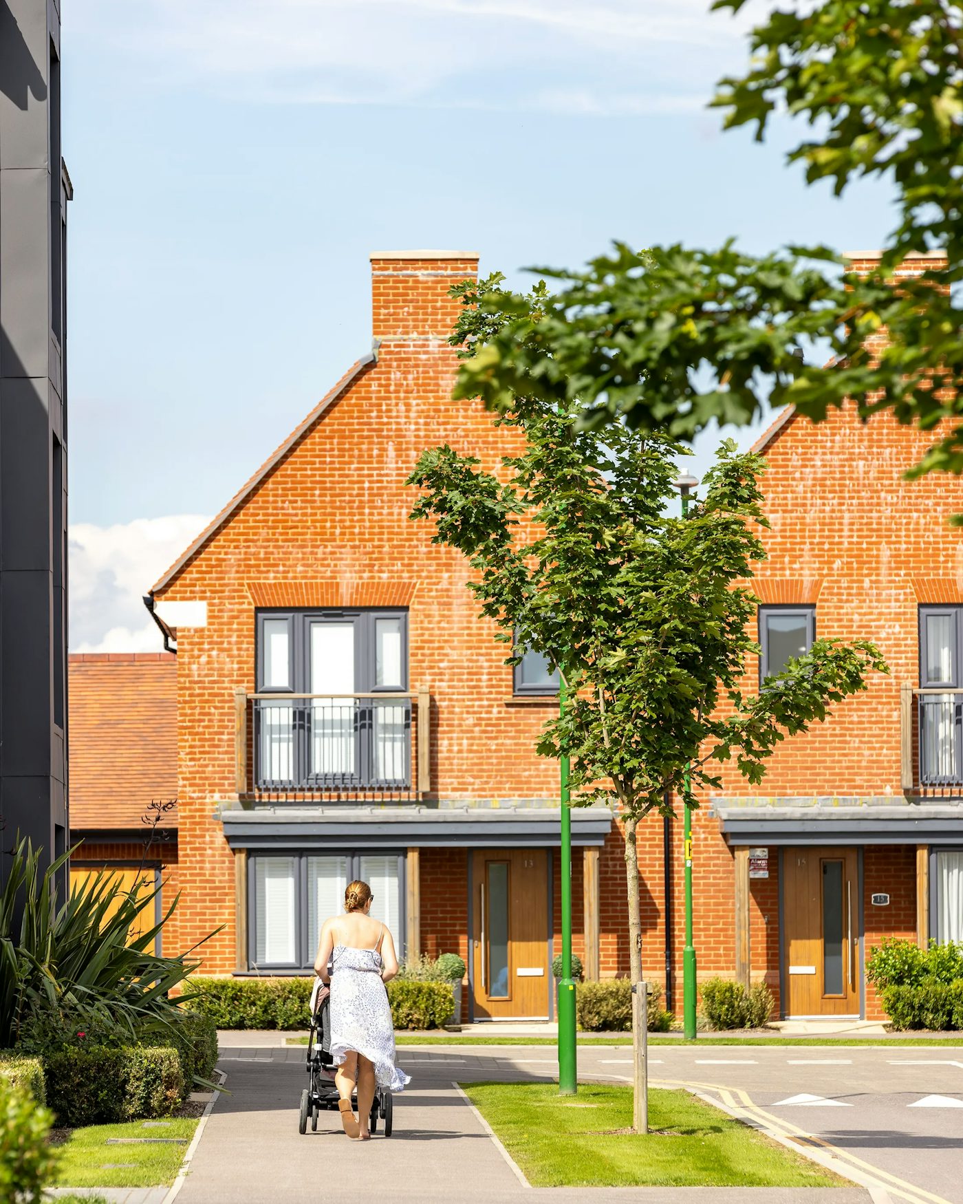 A person pushes a stroller along a paved path bordered by trees and greenery in front of a row of modern brick townhouses. The buildings feature large windows and compact balconies, with a bright sky and scattered clouds overhead, evoking a calm, family-friendly residential atmosphere.