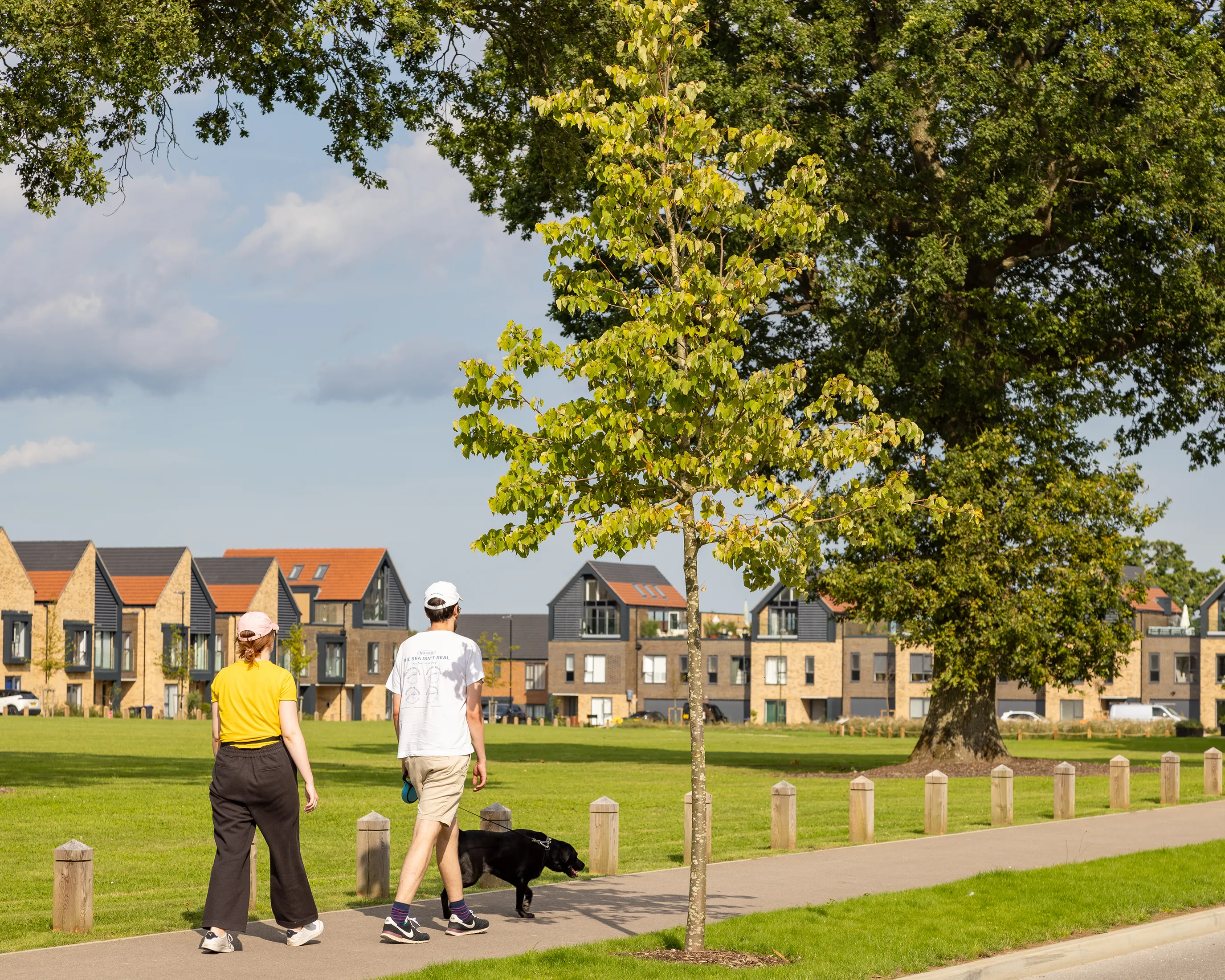 Two people walk a black dog along a wide, paved path bordered by trees and grass in a residential park. One wears a yellow top with dark trousers, the other a white shirt with light shorts; both wear caps. Behind them, modern brick and dark-panelled homes with red-tiled roofs line the horizon beneath a partly cloudy sky.