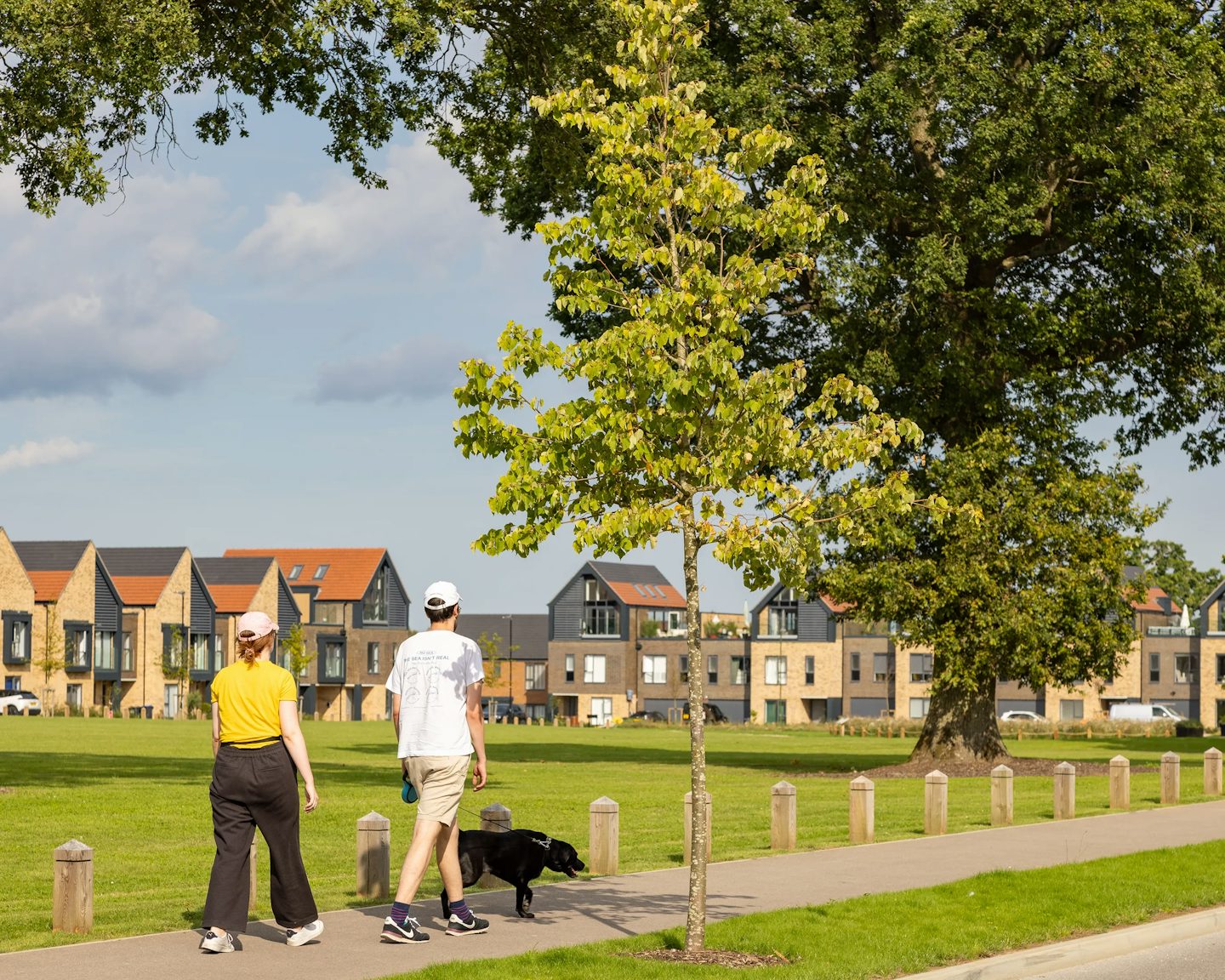 Two people walk a black dog along a wide, paved path bordered by trees and the village green at Woodgate. One wears a yellow top with dark trousers, the other a white shirt with light shorts; both wear caps. Behind them, modern brick and dark-panelled homes with red-tiled roofs line the horizon beneath a partly cloudy sky.