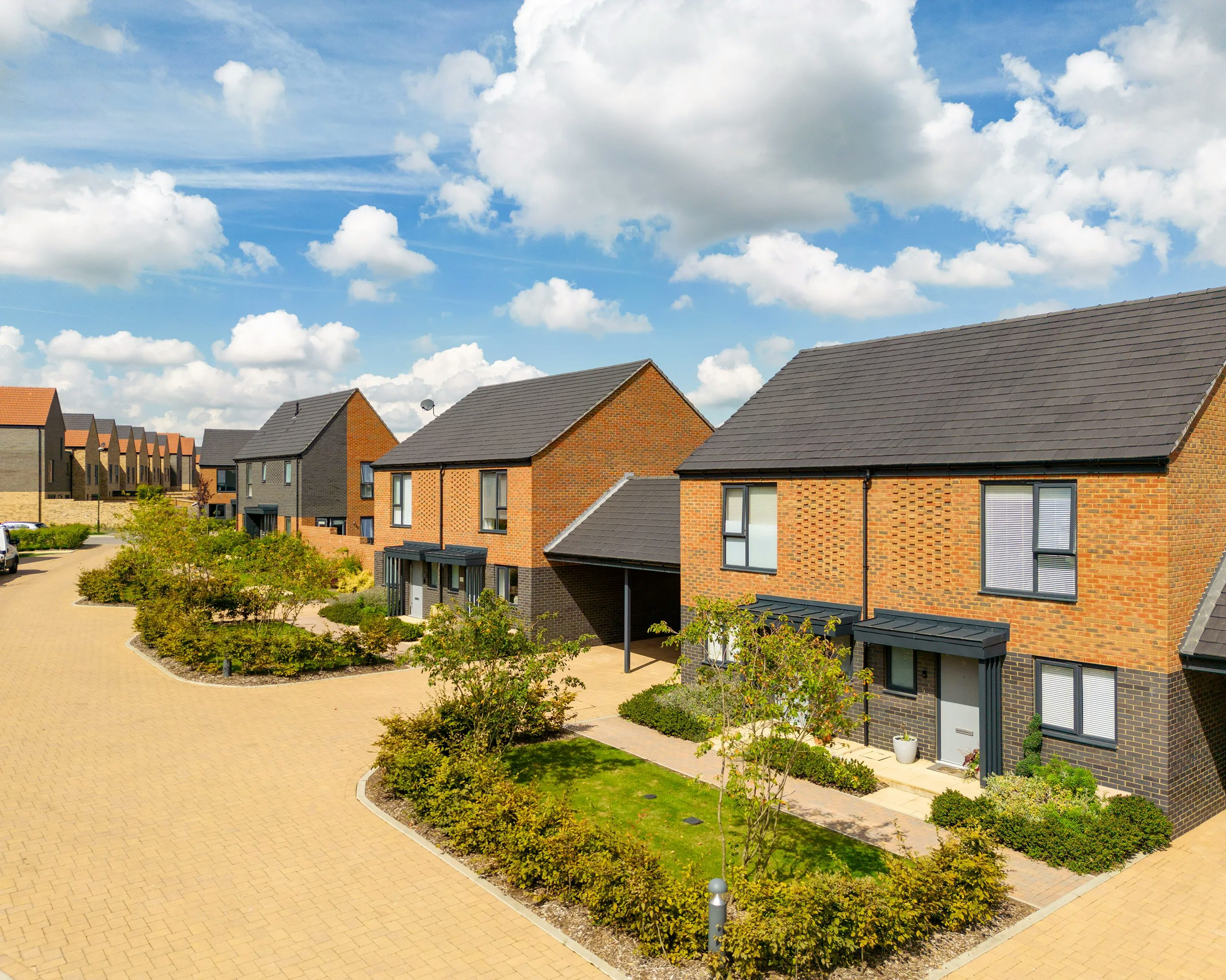 A modern residential street with two-story houses featuring brick and dark siding exteriors. Each home has large windows, well-kept front gardens, and landscaped borders with shrubs and small trees. The paved street is made of light-colored bricks and the sky is mostly clear, adding to the calm, welcoming atmosphere.
