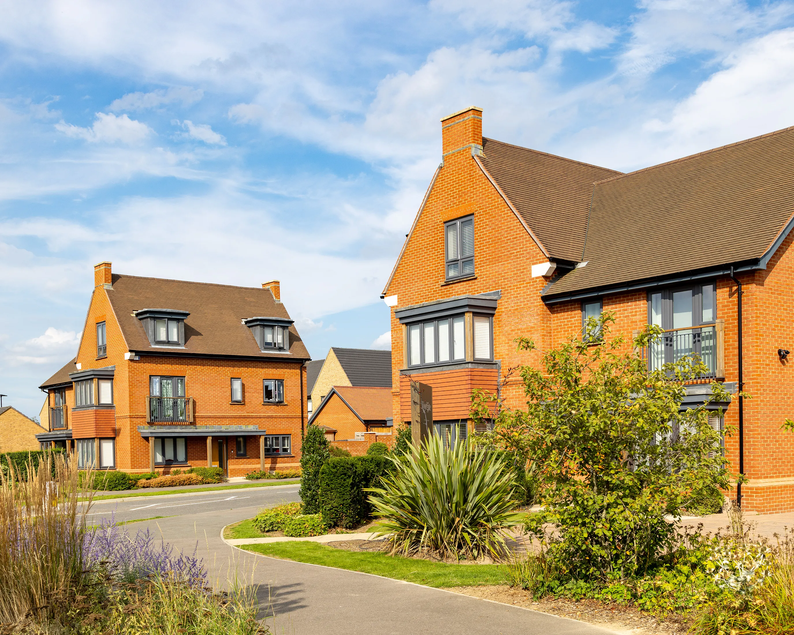 A residential street lined with modern, multi-storey brick houses featuring large windows, balconies with glass or metal railings, and steeply pitched roofs. The scene is bathed in sunlight beneath a vivid blue sky dotted with wispy clouds. Landscaped greenery, including shrubs, small trees, and a neatly trimmed lawn, frames the foreground, suggesting a well-kept and welcoming neighbourhood.