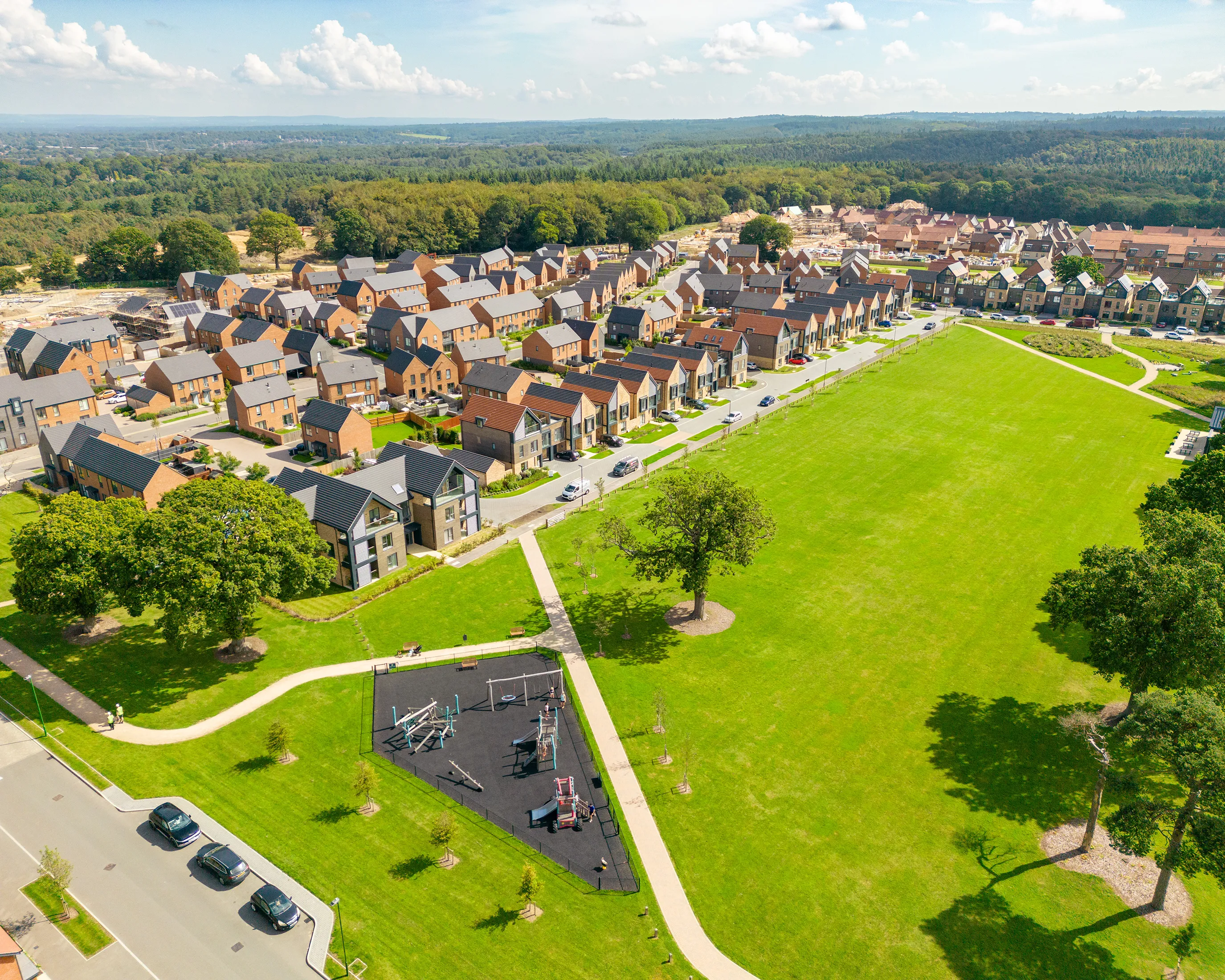 Aerial view of a planned residential area featuring rows of uniformly styled houses, a large green field occupying the center right, and a colorful playground in the bottom left corner. The layout highlights a balance between built structures and open communal space, with pathways connecting homes and recreational areas.