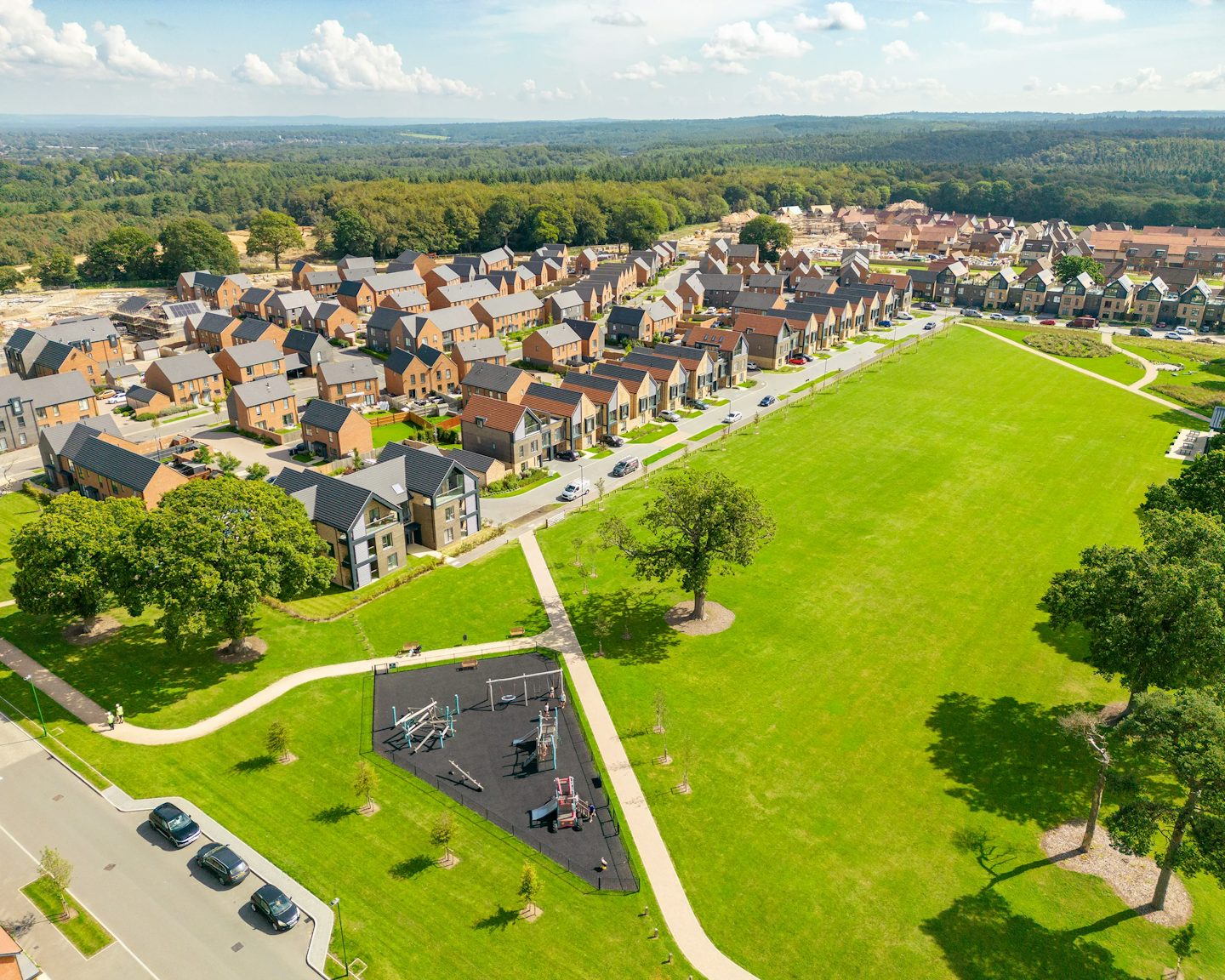 Aerial view of a planned residential area featuring rows of uniformly styled houses, a large green field occupying the center right, and a colorful playground in the bottom left corner. The layout highlights a balance between built structures and open communal space, with pathways connecting homes and recreational areas.