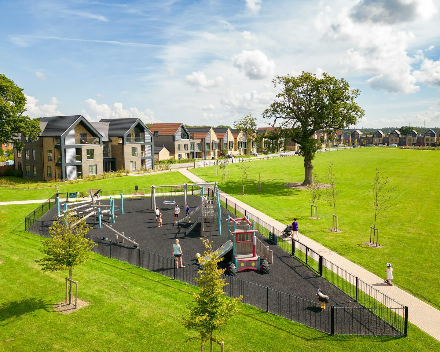 A fenced community playground with swings, slides, and climbing structures, set on a black safety surface. Children are actively playing, while a few adults walk along an adjacent paved path. Behind the play area, modern houses with pitched roofs line the horizon, and a grassy field with scattered trees extends into the distance beneath a partly cloudy sky.