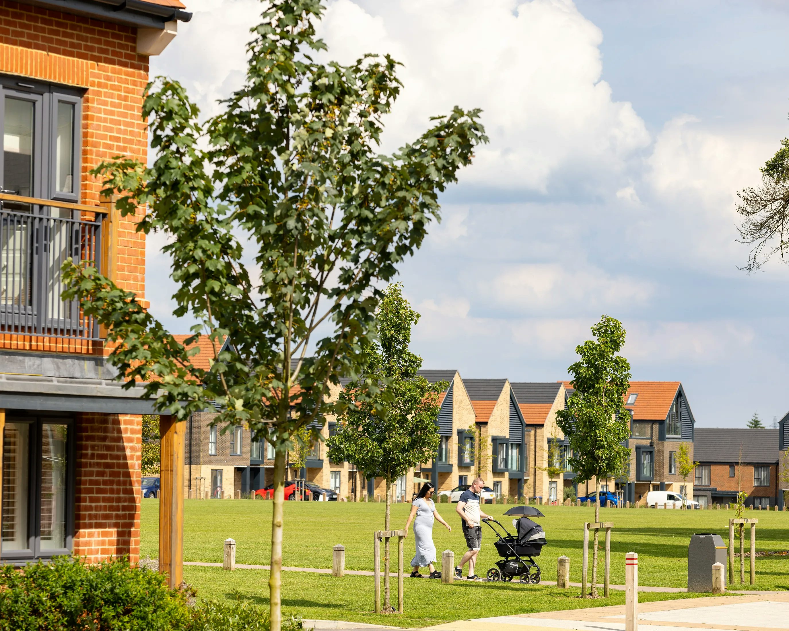 A tranquil suburban scene with modern houses lining a neatly trimmed green lawn. The homes feature a mix of brick and siding exteriors with large windows, framed by young trees. A few people walk along a central paved path, including one person pushing a stroller, reinforcing the family-friendly atmosphere. The sky is lightly overcast, suggesting mild weather.