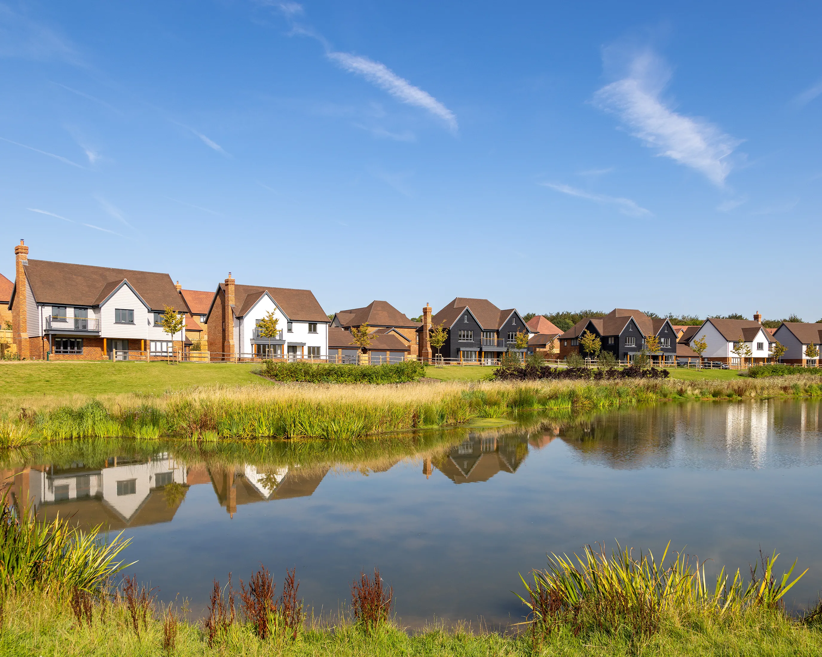A row of modern houses lines the far side of a calm pond, their brick and painted facades reflected in the still water. Reeds and lush greenery frame the foreground, while a clear blue sky enhances the scene’s peaceful, residential charm.
