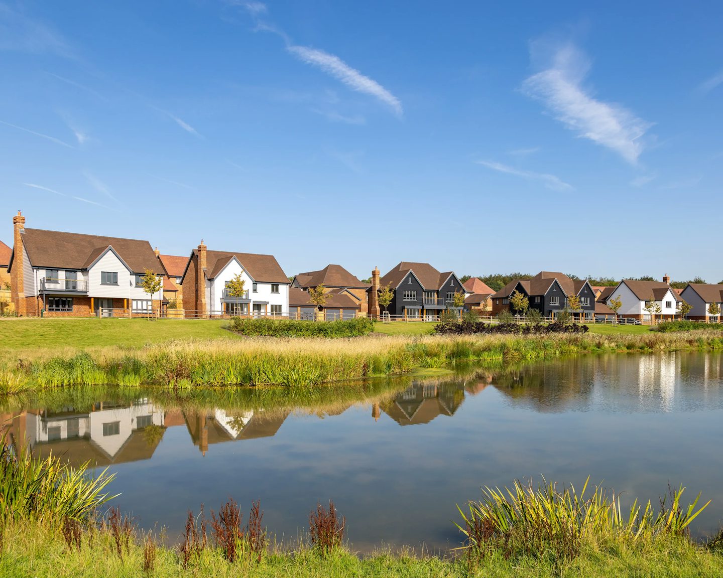 A row of modern houses lines the far side of a calm pond, their brick and painted facades reflected in the still water. Reeds and lush greenery frame the foreground, while a clear blue sky enhances the scene’s peaceful, residential charm.