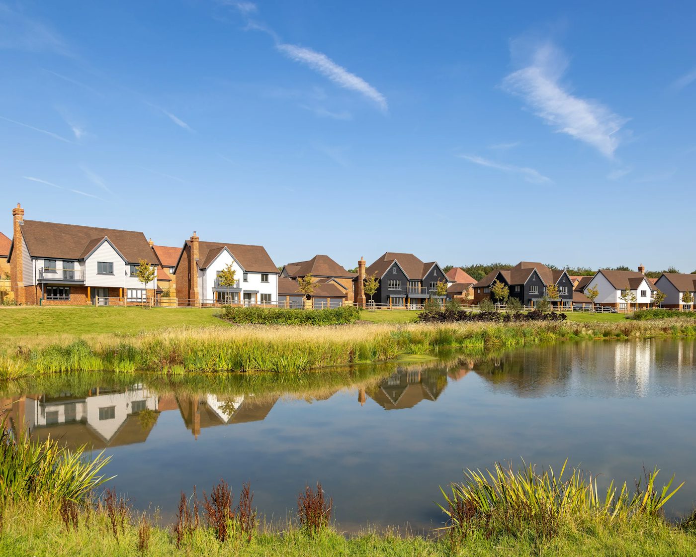 A row of modern houses lines the far side of a calm pond, their brick and painted facades reflected in the still water. Reeds and lush greenery frame the foreground, while a clear blue sky enhances the scene’s peaceful, residential charm.