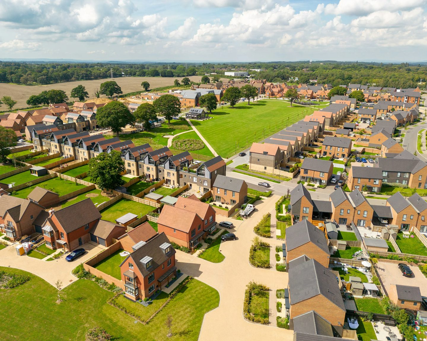 Aerial view of Woodgate featuring rows of detached and semi-detached houses with pitched roofs, interspersed with green lawns and tree-lined streets. A large rectangular village green sits at the centre, bordered by pathways and surrounded by homes, creating a sense of openness and community within the residential layout.