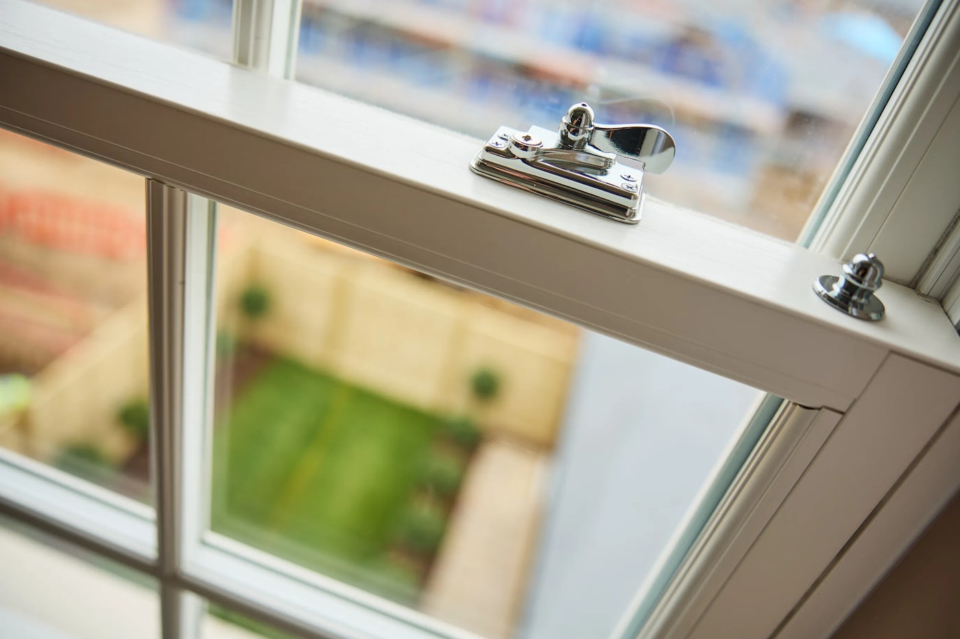 Close-up of a partially open window with a silver latch in the locked position, overlooking a tidy garden with a green lawn, small shrubs, and a wooden fence.