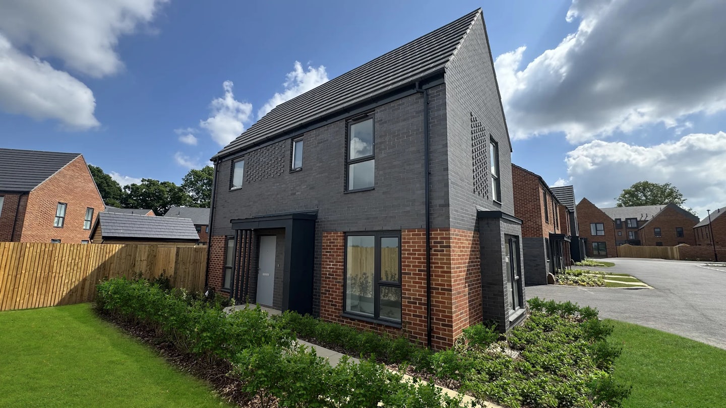 A modern two-story house with a dark gray and red brick exterior, featuring large windows, a small manicured garden with green shrubs, and a neatly trimmed lawn. Set in a residential neighbourhood with similar houses in the background, under a partly cloudy sky.