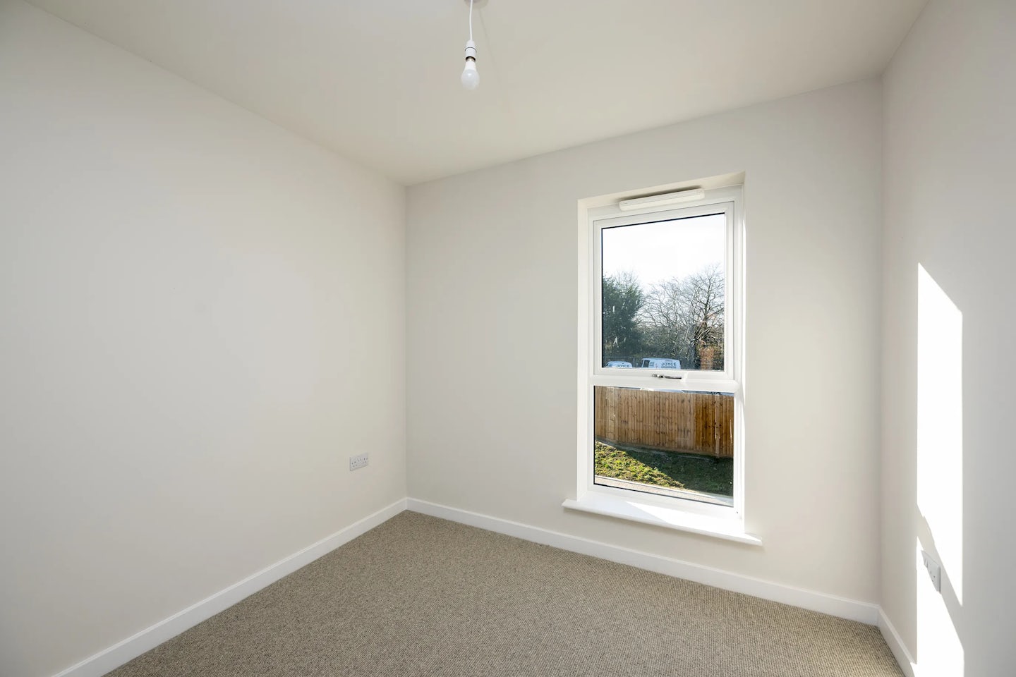 A small, unfurnished room with white walls and a beige carpeted floor. A single window on the far wall lets in natural light and reveals a view of a wooden fence, green grass, and leafy trees. A bare light bulb hangs from the ceiling, emphasizing the simplicity of the space.