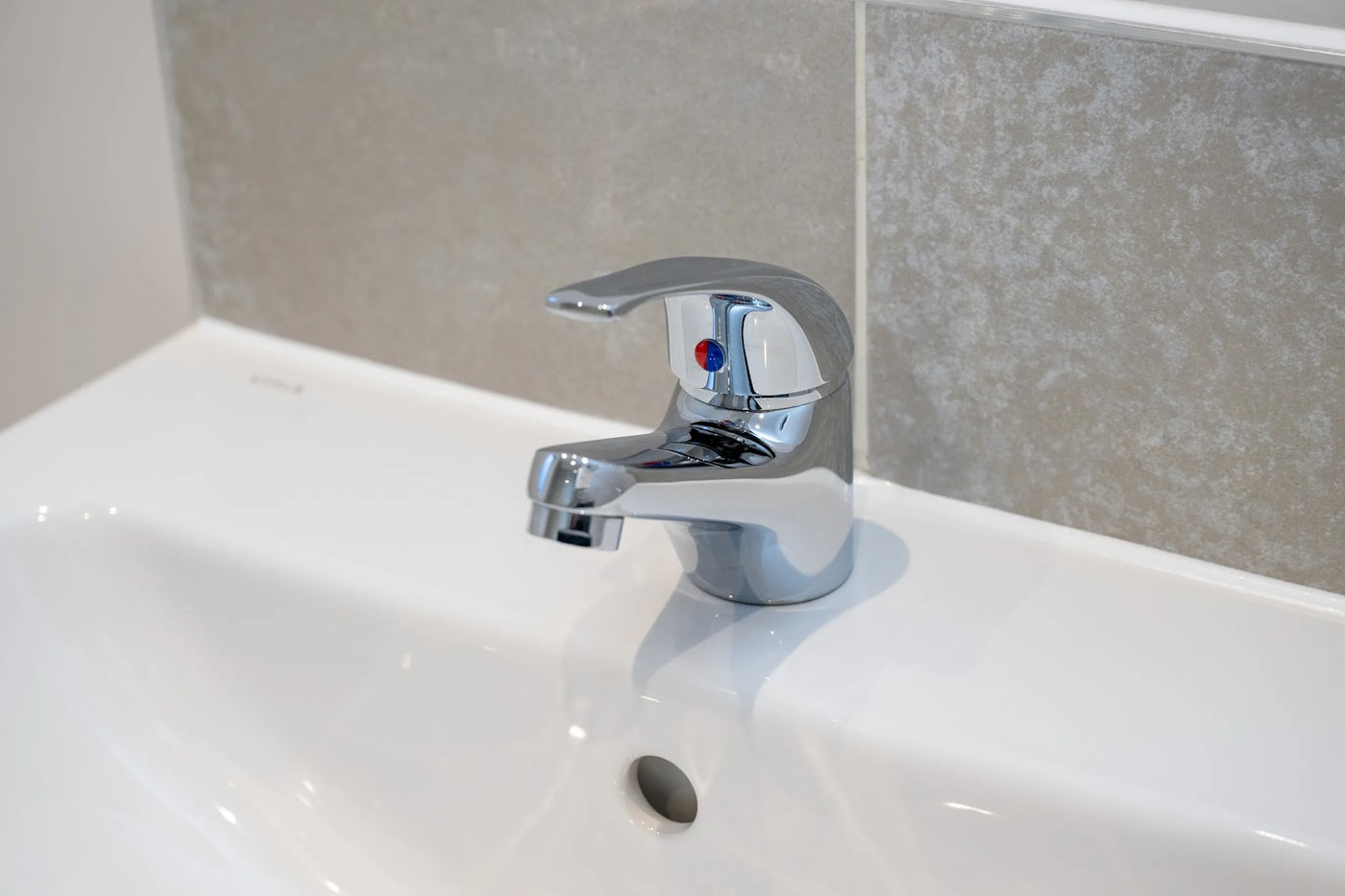 Close-up of a modern chrome bathroom faucet with a single lever handle, mounted on a white sink. A small circular indicator with red and blue markings sits on the handle, denoting hot and cold water. The grey tiled wall in the background adds a neutral, contemporary backdrop.