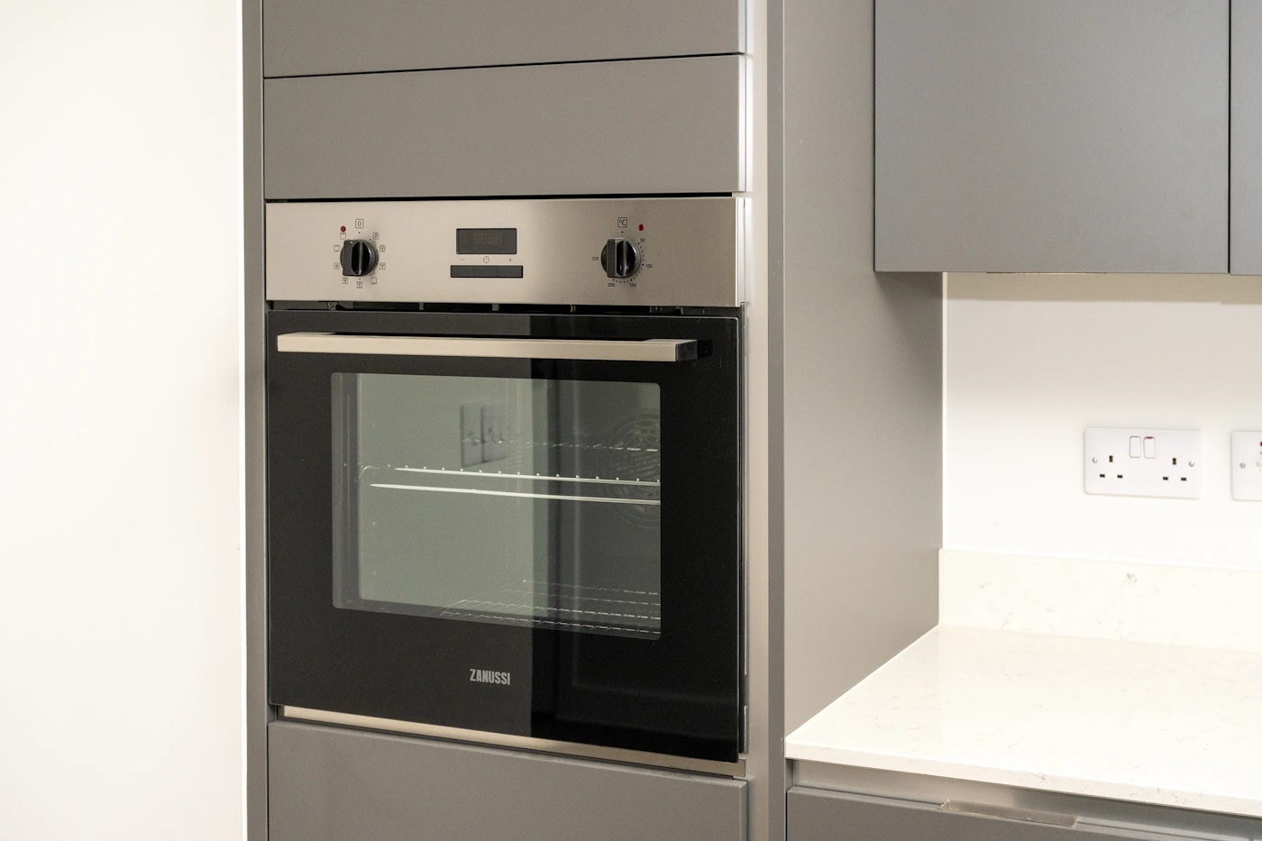 A built-in stainless steel Zanussi oven with two control knobs and a digital display is set into white cabinetry. To the right, a smooth white countertop runs alongside a wall outlet, contributing to a clean, modern kitchen setup.