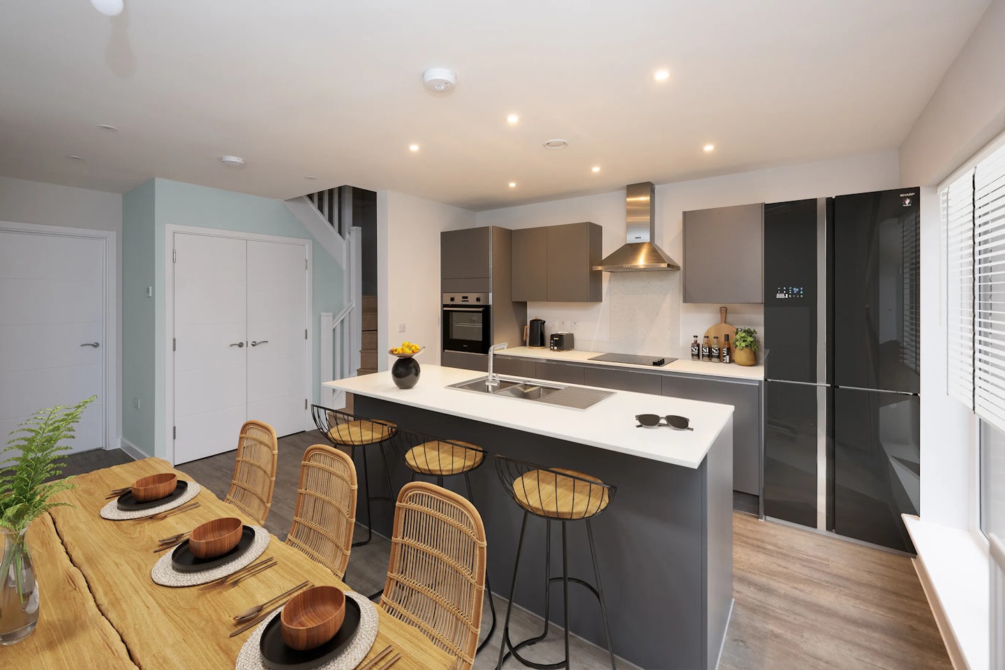 Modern kitchen and dining area with grey cabinetry and natural wood accents. A white-topped island with a built-in sink and stovetop sits centrally, paired with four wicker bar stools. Behind it, matte grey cabinets, a stainless steel oven, and a matching range hood frame the workspace. To the right, a large black refrigerator contrasts the pale flooring. The adjacent dining area features a wooden table set for four, with coordinated brown bowls and plates. A staircase and closed door are visible in the softly lit background, all framed by recessed ceiling lights.