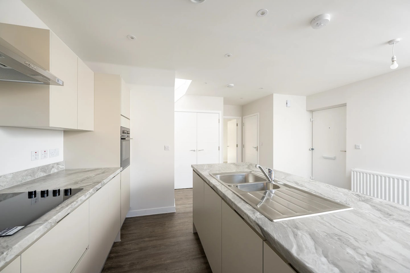 A contemporary kitchen with light cabinetry, marble countertops, and stainless steel appliances. A central island features a sink and drying rack, while dark wood flooring contrasts with the light-toned walls. The space is brightly lit by recessed lighting and a skylight, enhancing the clean, minimalist design.