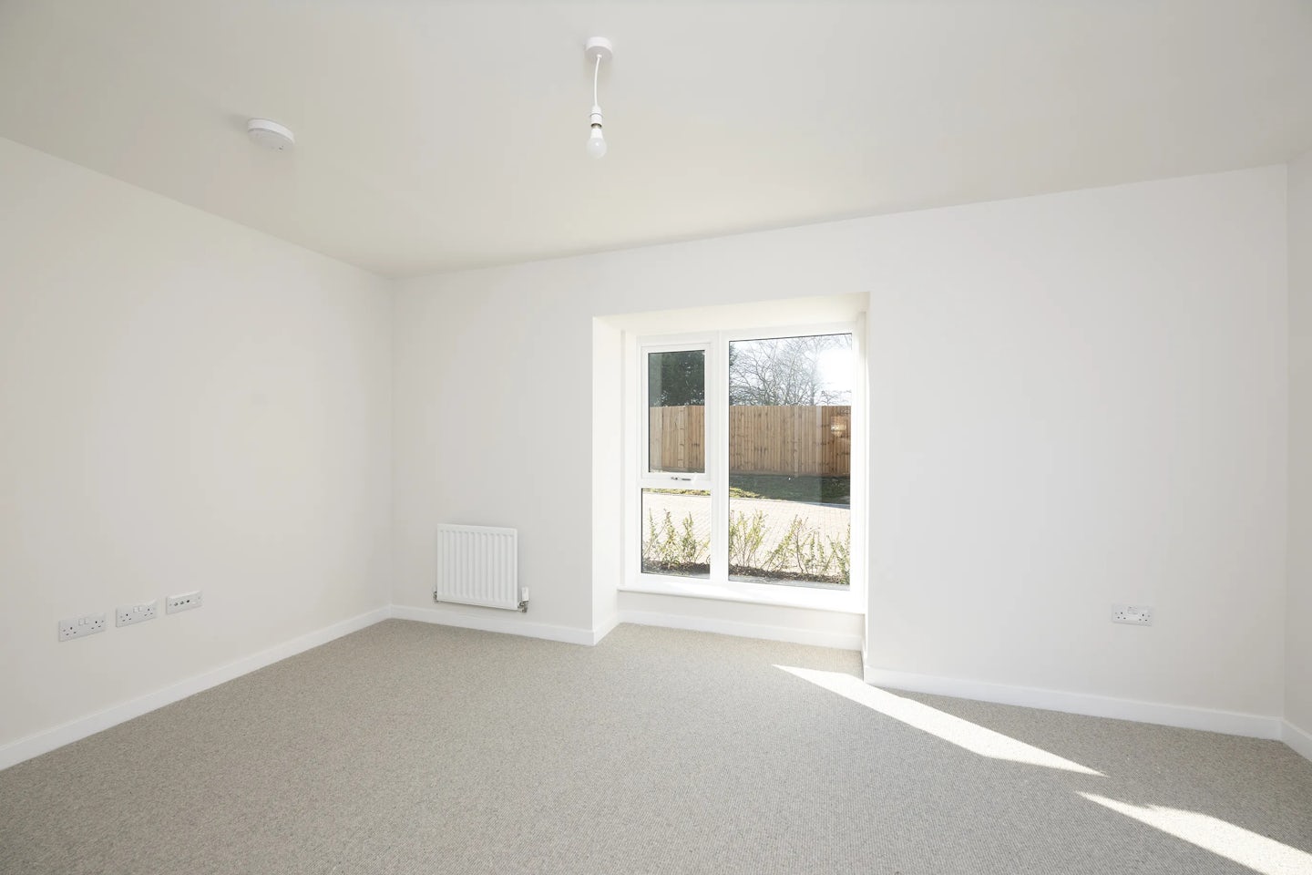 A bright, unfurnished room with white walls and a beige carpet. A wide window on the right lets in soft natural light and offers a view of a garden bordered by a wooden fence. A radiator sits below the window, and a simple ceiling light fixture adds to the room's clean, neutral atmosphere
