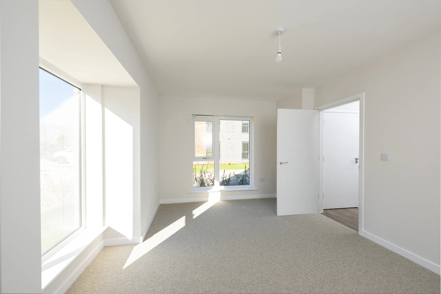 Unfurnished, sunlit room with beige carpet and white walls, featuring two large windows and an open door leading to a darker-floored area.