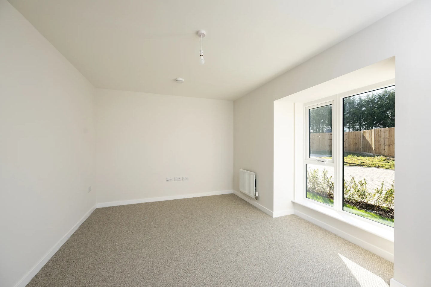 A bright, unfurnished room with white walls and a beige carpet. A wide window on the right lets in soft natural light and offers a view of a garden bordered by a wooden fence. A radiator sits below the window, and a simple ceiling light fixture adds to the room's clean, neutral atmosphere