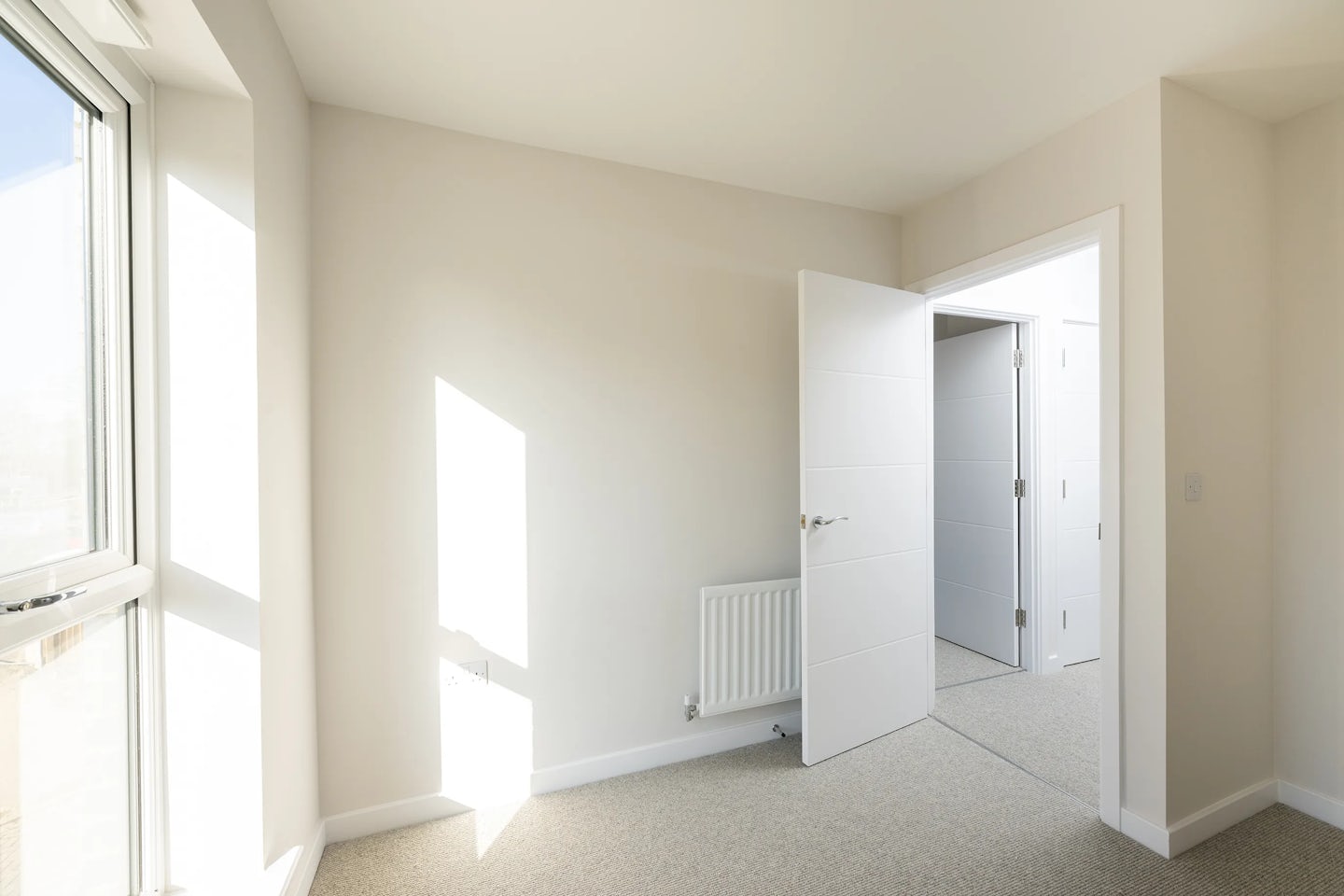 A bright, minimalistic room with beige walls and carpet, lit by natural daylight streaming through a large window on the left. The sunlight casts a bold rectangular shadow on the far wall above a white radiator. An open white door reveals a glimpse of a similarly styled adjoining space. The overall feel is calm, uncluttered, and softly illuminated.