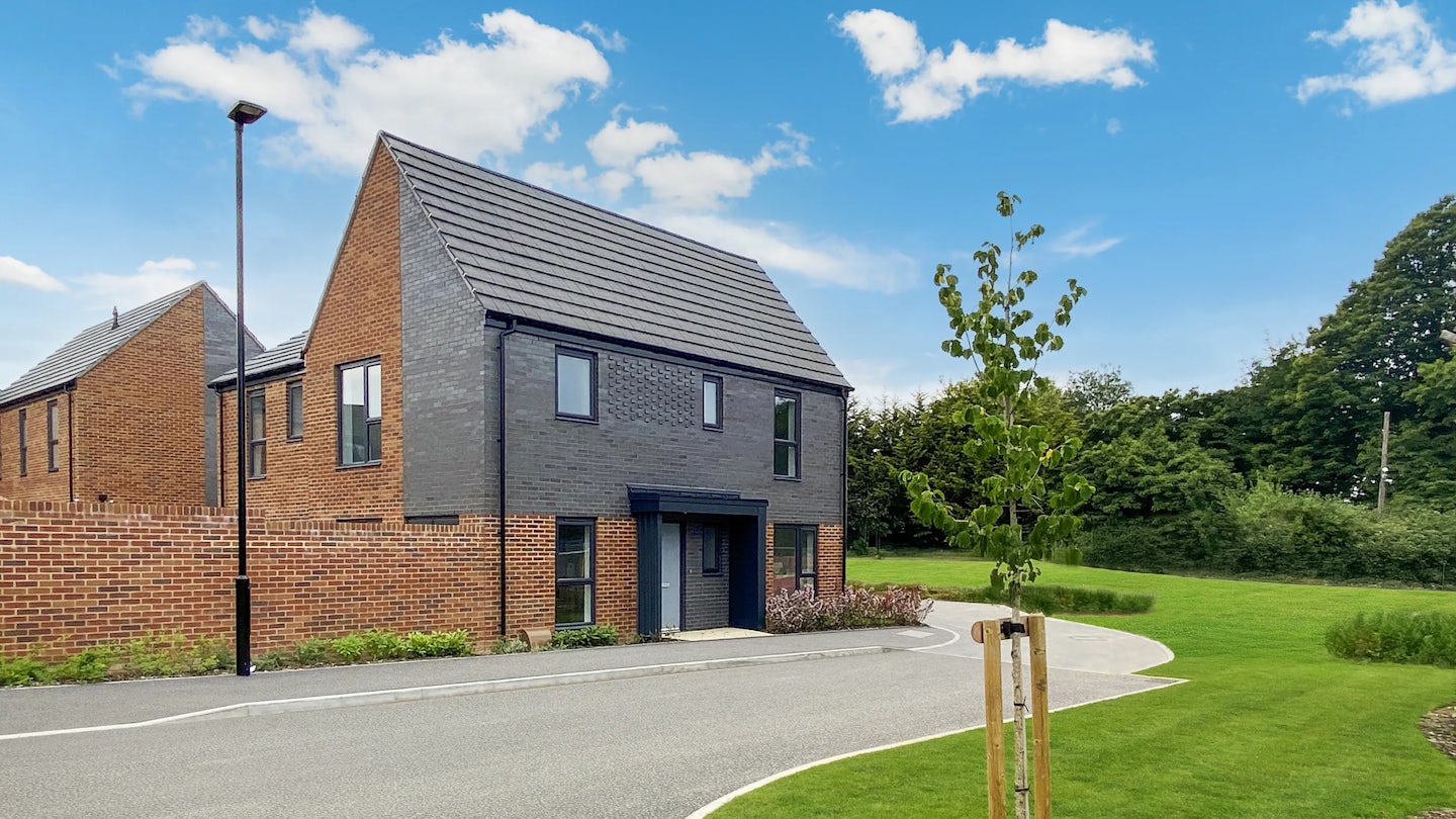 A modern two-story house with dark grey and red brick cladding, featuring a pitched grey-tiled roof and multiple rectangular windows. A young tree, supported by wooden stakes, stands in the neat front yard near a curved residential road and streetlamp. A similar house and green foliage appear in the background under a lightly clouded sky.