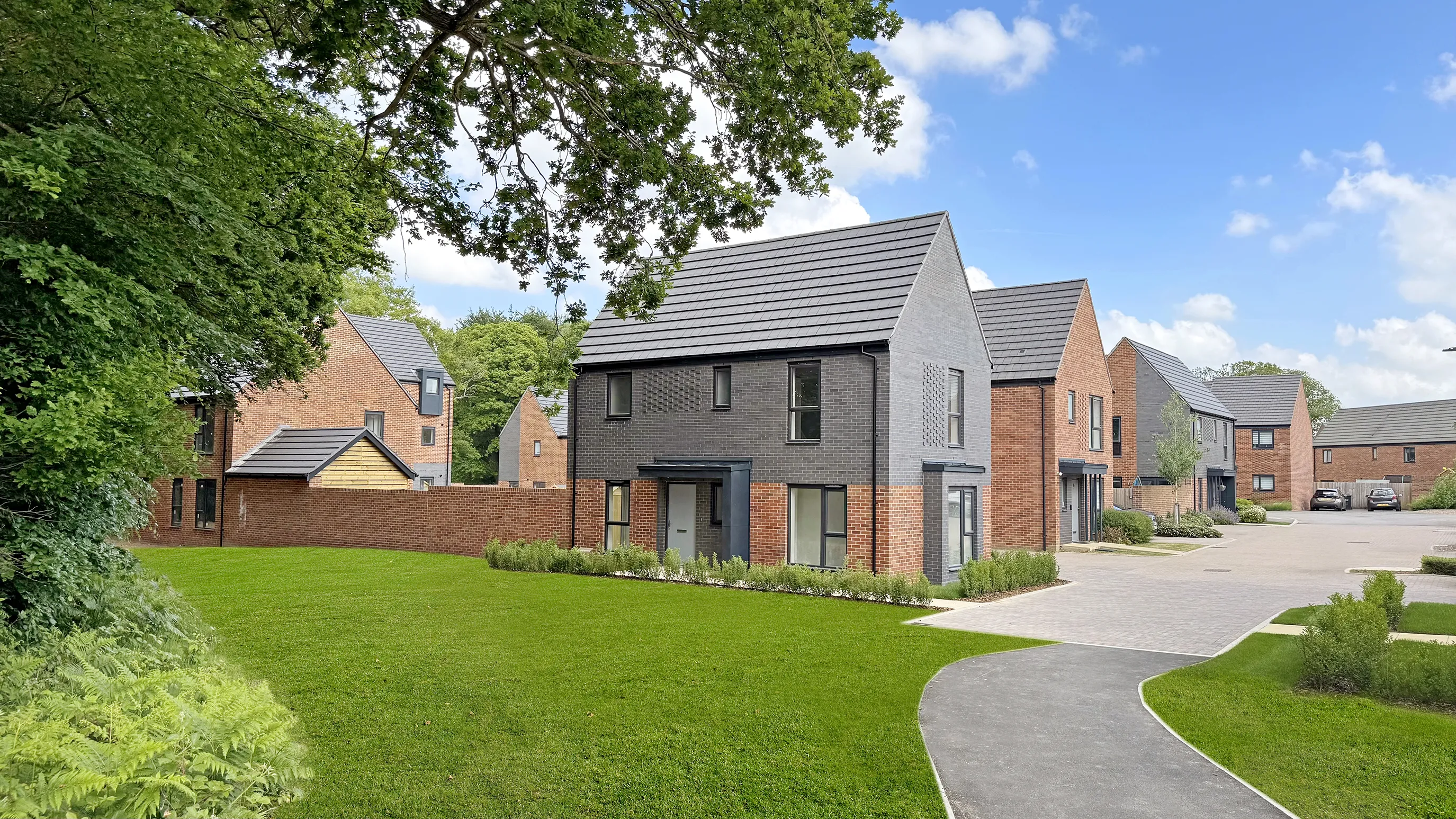 A modern suburban street featuring several newly built houses with a mix of red brick and dark grey cladding. The homes are set along a paved walkway and surrounded by neatly trimmed grass, young trees, and small shrubs. A driveway in the background holds a few parked cars. The sky is mostly clear with a few scattered clouds, suggesting mild weather.
