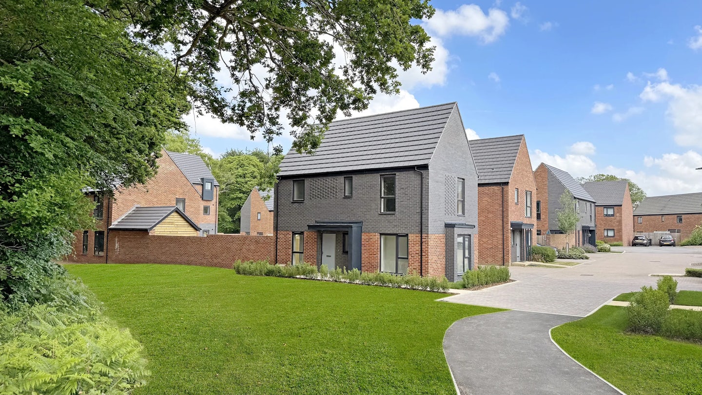 A modern suburban street featuring several newly built houses with a mix of red brick and dark grey cladding. The homes are set along a paved walkway and surrounded by neatly trimmed grass, young trees, and small shrubs. A driveway in the background holds a few parked cars. The sky is mostly clear with a few scattered clouds, suggesting mild weather.