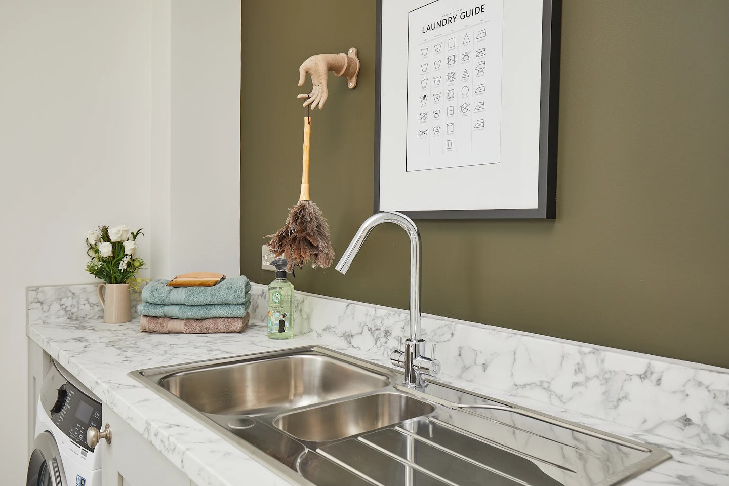 A compact laundry area featuring a marble countertop with a stainless steel sink. On the countertop are neatly folded white towels, a bottle of detergent, and a small vase with flowers. Above the sink, a framed “Laundry Guide” displays various laundry care symbols. A feather duster hangs on the wall nearby, adding a practical touch to the space.