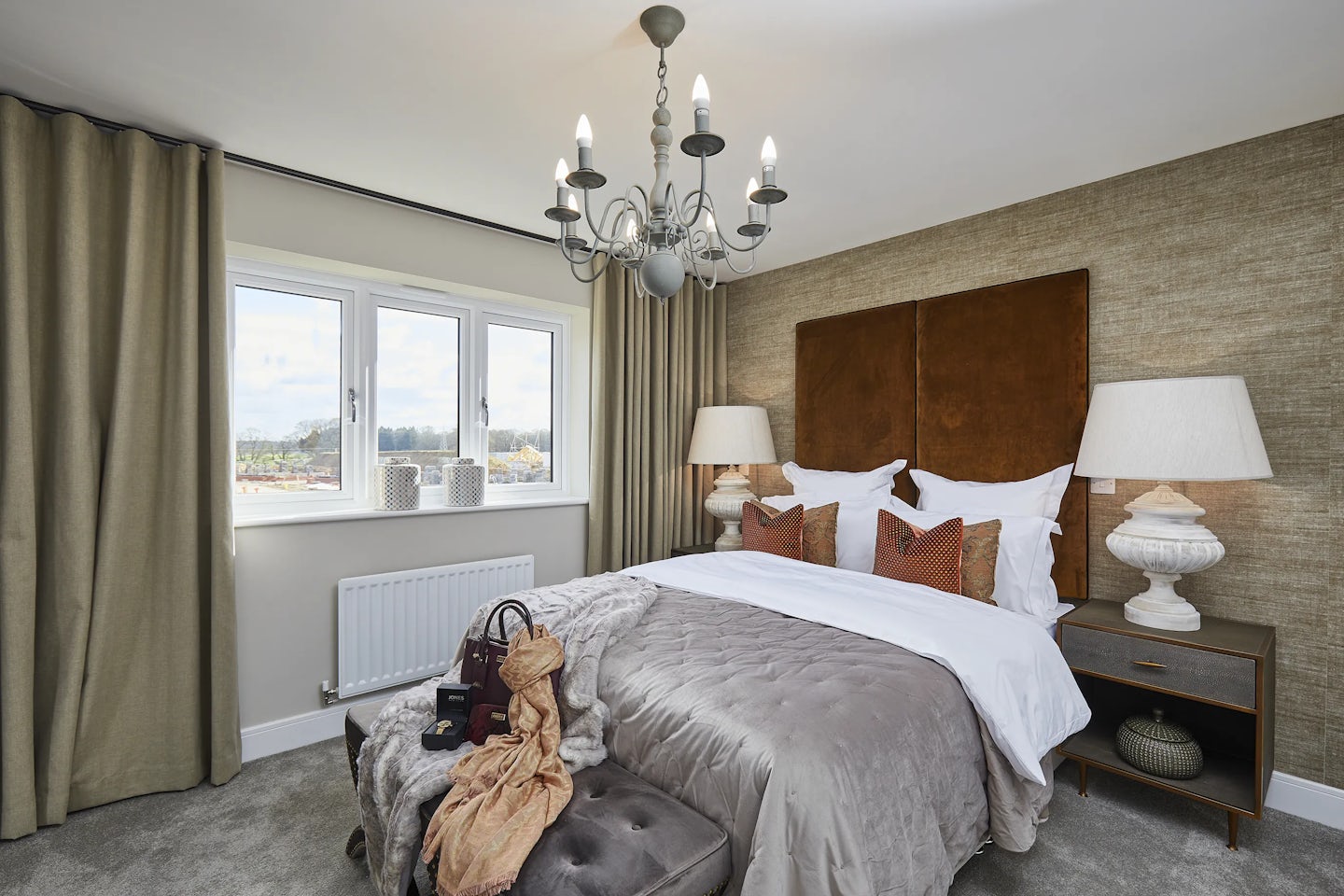 A softly lit bedroom with a double bed dressed in white sheets and a gray comforter, accented by large brown pillows. A tufted bench at the foot of the bed holds a tan handbag, a patterned scarf, and a closed book. Matching bedside tables with lamps flank the bed, and a chandelier hangs overhead. A window with neutral curtains reveals a daylight view, with decorative items on the sill and a white radiator below. The walls are painted in warm, neutral tones, creating a cozy and elegant atmosphere.
