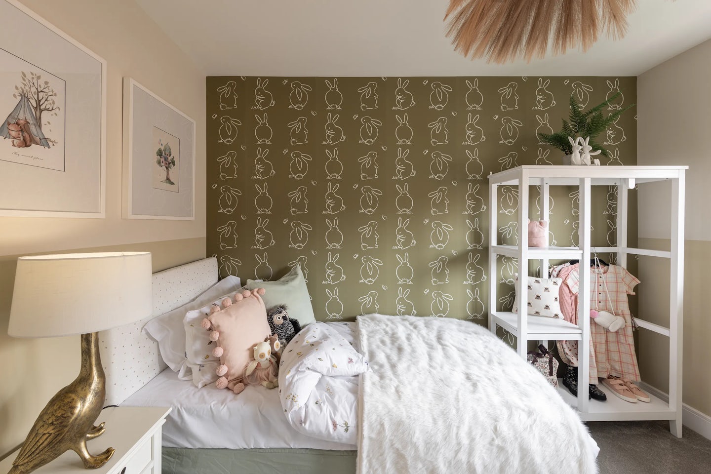 A softly lit children’s bedroom with a green accent wall patterned with white bunny outlines. A white-framed bed is dressed in pastel bedding and playful pillows. Beside it, a gold bird-shaped lamp rests on a nightstand. A white shelving unit displays clothes and decor, while framed prints and a sculptural ceiling light add whimsical touches.