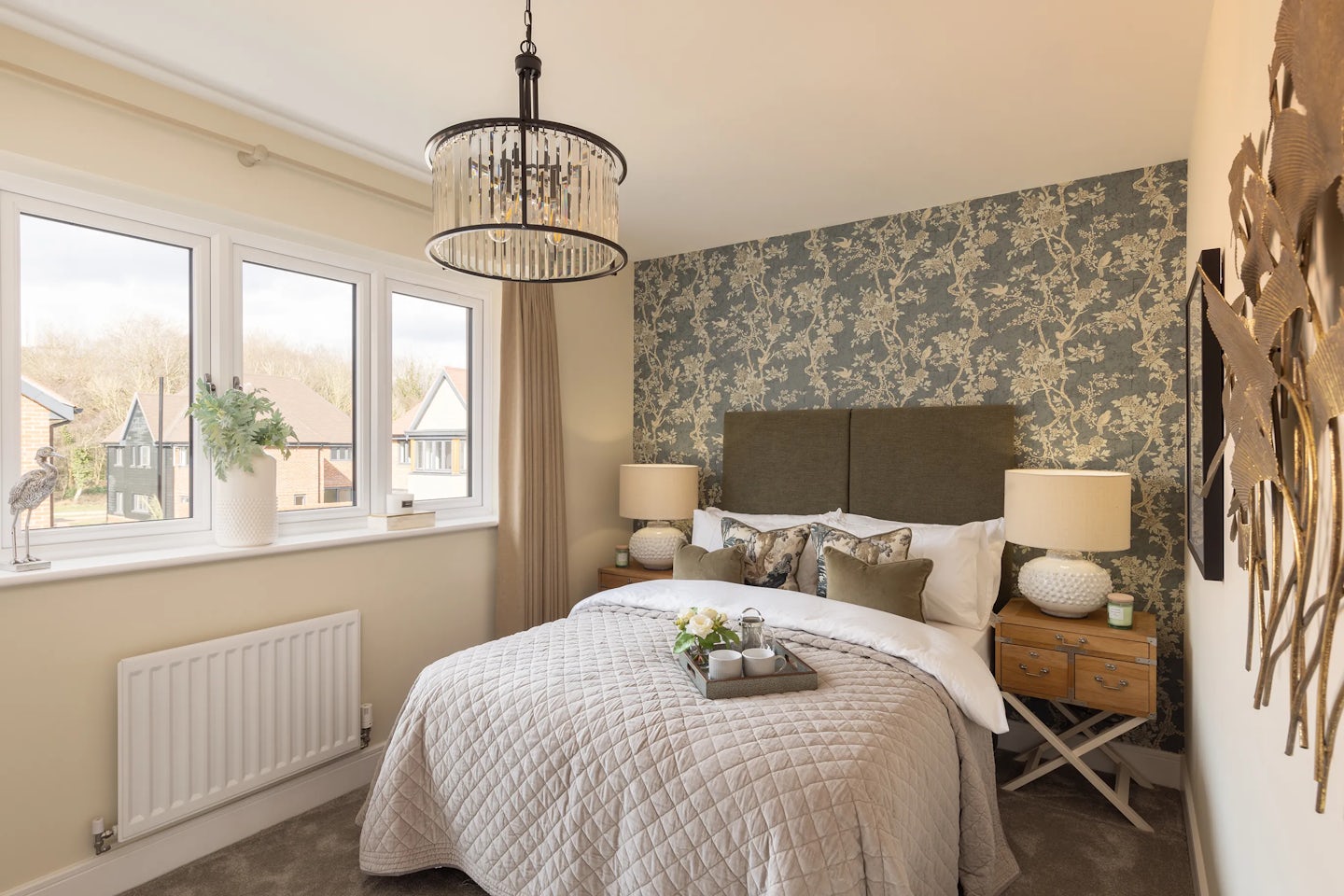 A bright, cozy bedroom with floral wallpaper behind a bed topped with a quilted bedspread. Two bedside tables with lamps flank the bed, and a chandelier hangs overhead. Sunlight streams through a large window, revealing rooftops and trees outside.