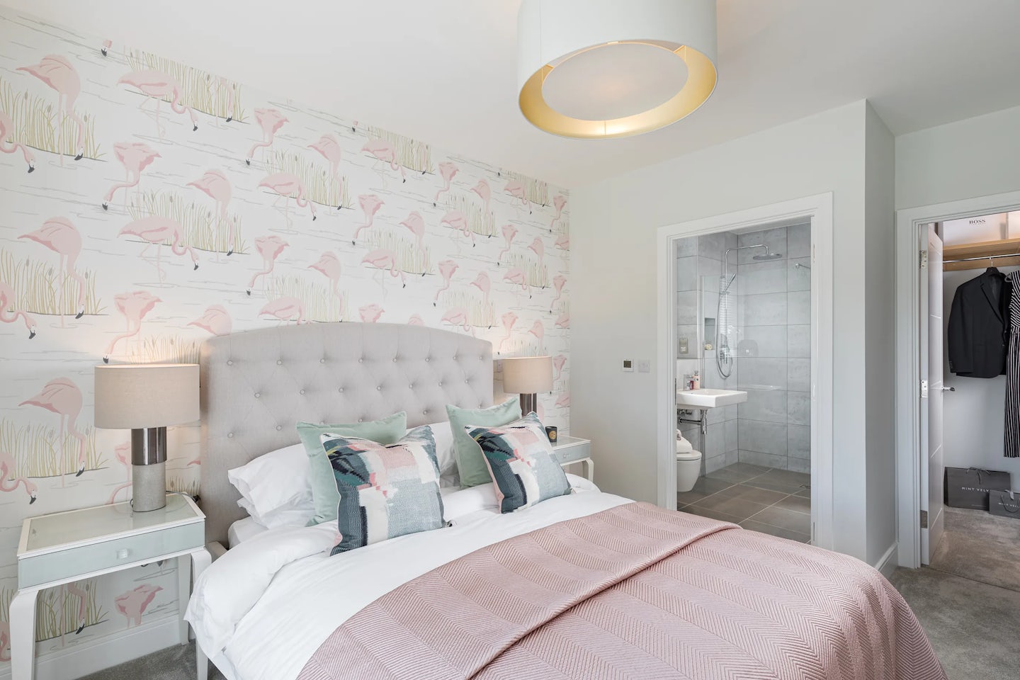 A softly lit bedroom with flamingo-patterned wallpaper and matching flamingo cushions on a bed with a tufted headboard. Two bedside tables with lamps sit on either side. An open doorway reveals a compact en-suite bathroom, and another leads to a closet with hanging clothes. Pale pinks and creams create a calm, coordinated atmosphere.