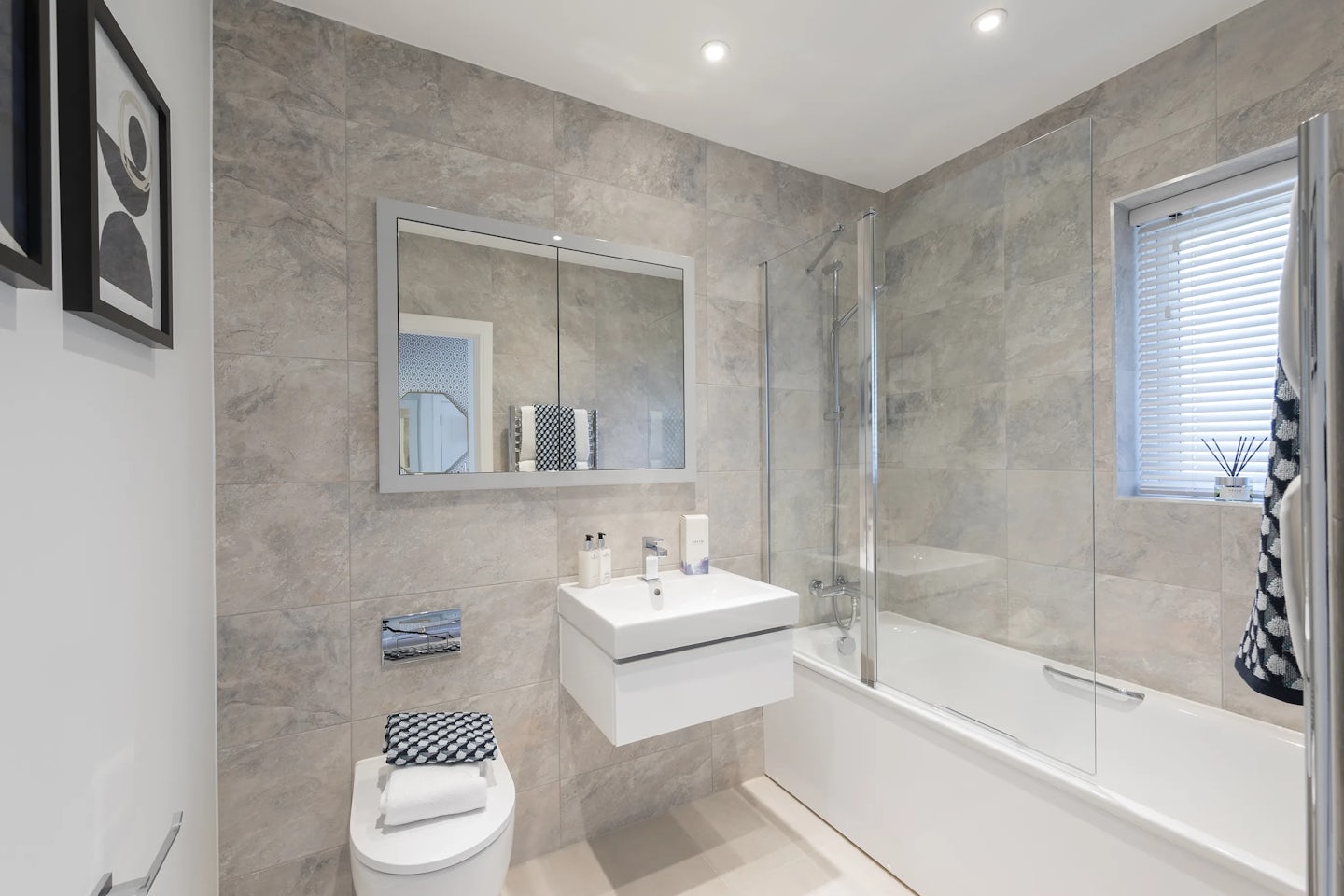 A contemporary bathroom featuring a wall-mounted sink with toiletries, a toilet, and a bathtub with a glass shower screen. Light grey stone-pattern tiles cover the walls, while a window with blinds adds natural light. Two framed pictures hang beside the sink, and the space is lit with recessed ceiling lights.
