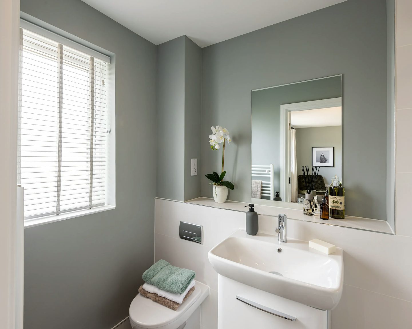 A modern, light-filled bathroom featuring a white sink with chrome fixtures, a soap dispenser, and a bar of soap on the counter. To the left sits a toilet topped with neatly folded towels. A potted plant with white flowers rests on the ledge above, while a large mirror and a window with white blinds reflect the clean, minimalist design.