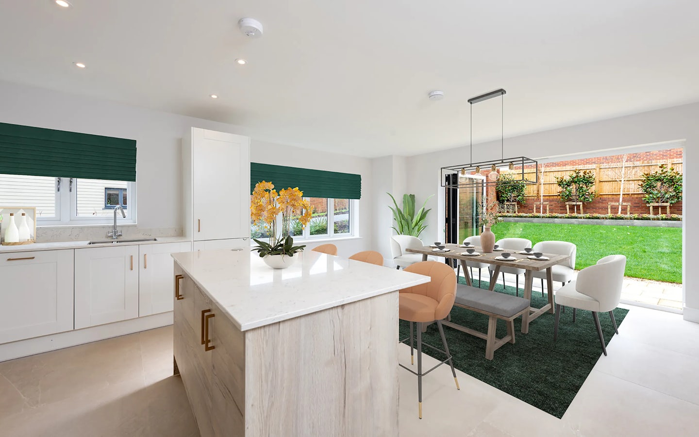 A contemporary open-plan kitchen and dining area featuring white cabinetry, a marble island with gold handles, and green blinds over twin windows above the sink. A six-seat dining table, set atop a muted green rug, blends beige and white chairs beneath a modern chandelier. Large sliding glass doors reveal a neatly kept backyard with a lush lawn, wooden fencing, and leafy border plantings, inviting ample natural light into the room.