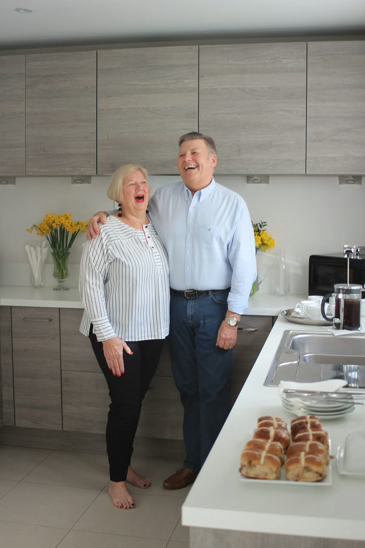 Two people stand closely together in a modern kitchen with light wood cabinets, white countertops, and stainless steel appliances. A vase of yellow flowers, a coffee maker, and a plate of hot cross buns sit on the counter, evoking a warm, inviting atmosphere.