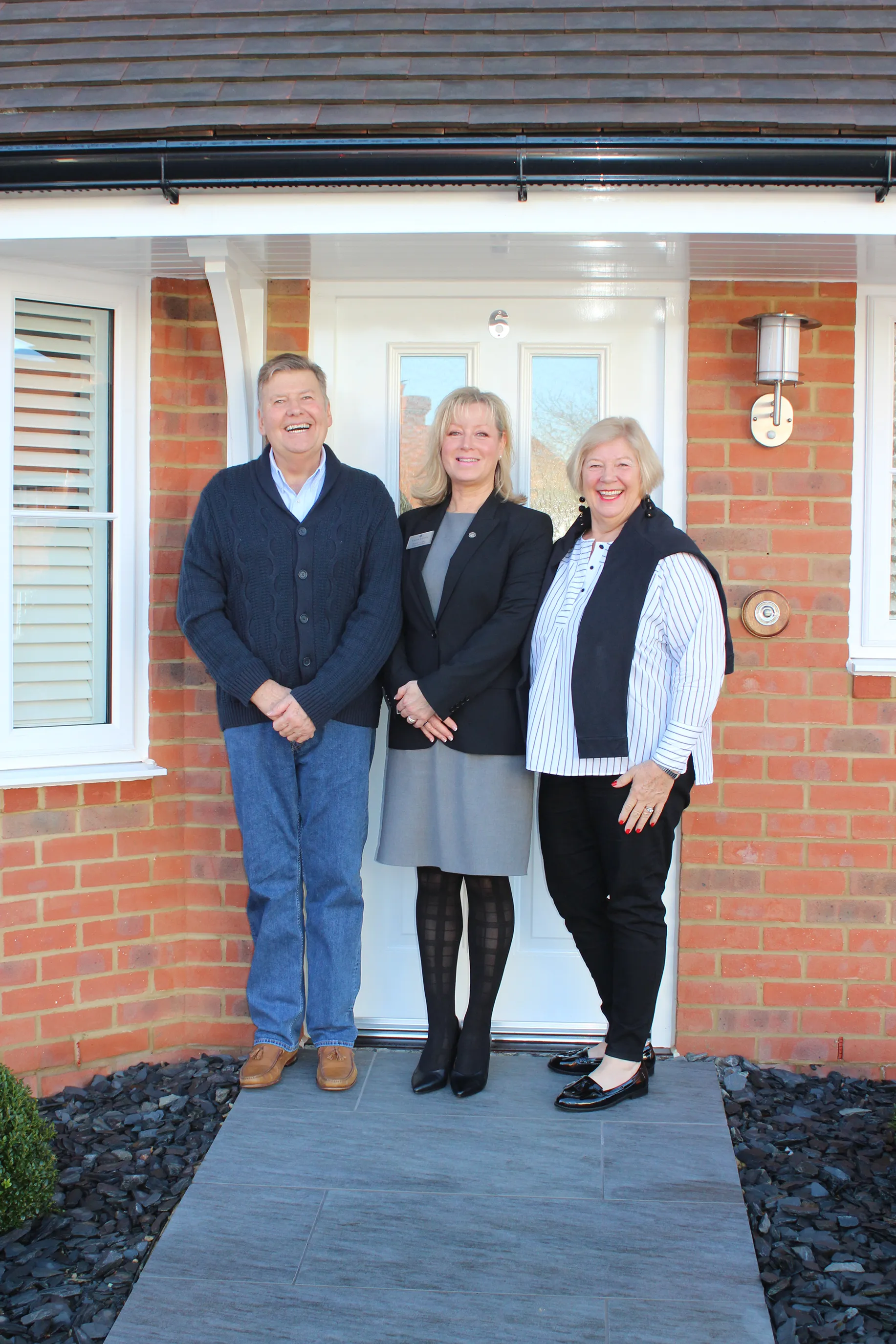 Three people standing at the entrance of a brick house. The entrance features a white door with glass panels and a small window with white shutters on the left. A gray doormat lies underfoot, and a landscaped corner with black stones and a green bush appears in the bottom left. The person in the middle wears a gray dress with a black blazer; the person on the left wears a blue sweater with jeans and brown shoes; the person on the right wears a striped shirt, black vest, black trousers, and black shoes.