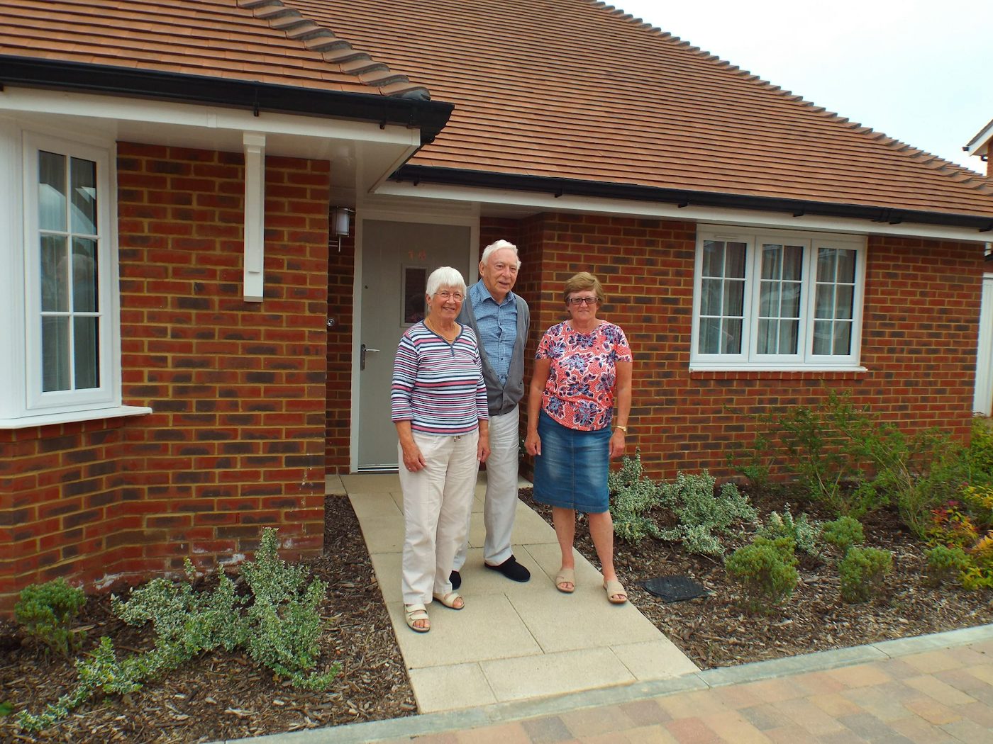 Three people stand outside a brick house with a tiled roof, gathered on a paved walkway leading to a white door. The home features white-trimmed windows and is framed by a small front garden with leafy shrubs and plants, suggesting a lived-in, welcoming atmosphere.