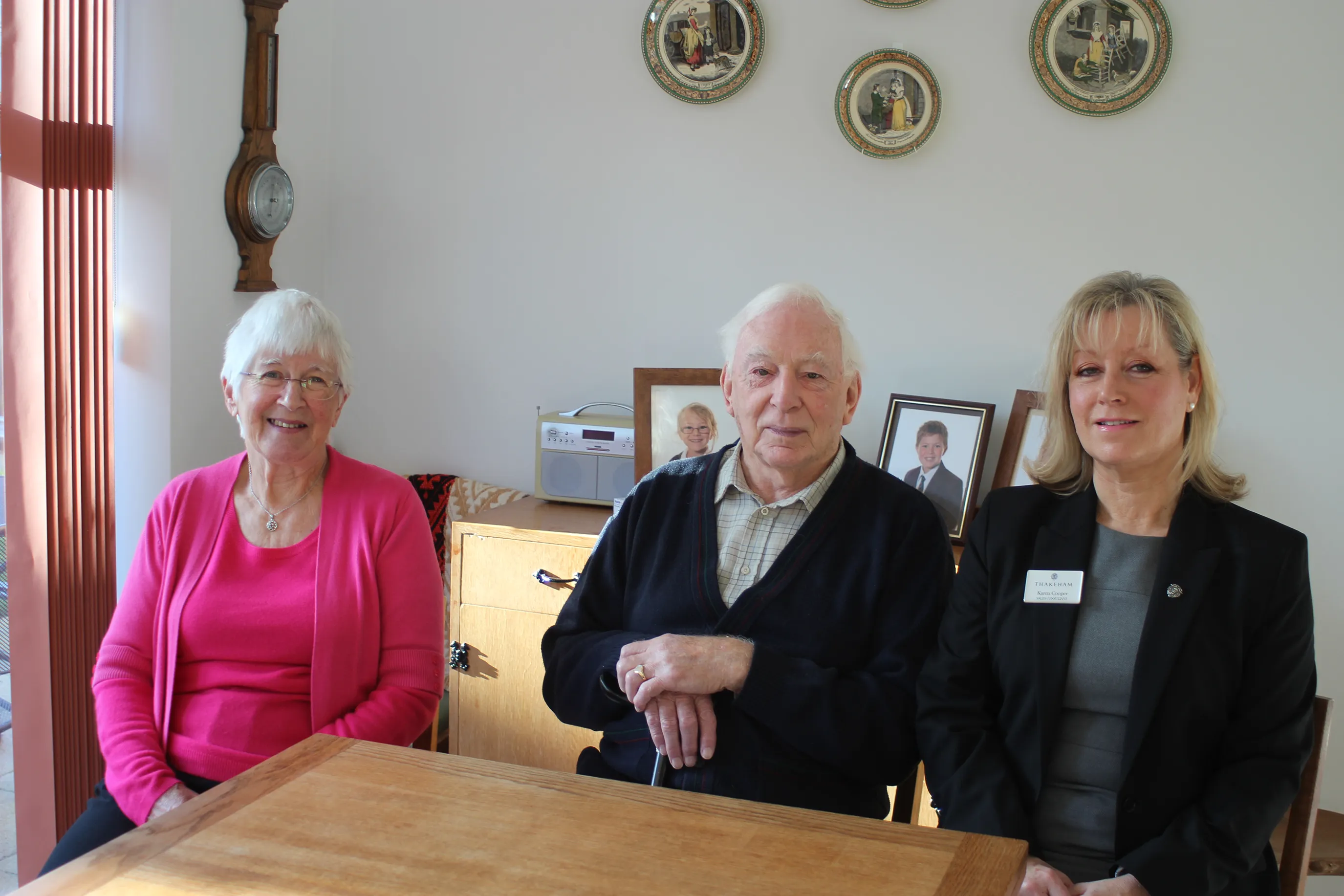 A wooden table with three people seated, engaged in conversation. Behind them is a wooden cabinet topped with framed photographs and flanked by a wall-mounted clock. Above the cabinet, four decorative plates are arranged in a row on a neutral-toned wall. All three individuals are dressed formally; one wears a visible name tag.