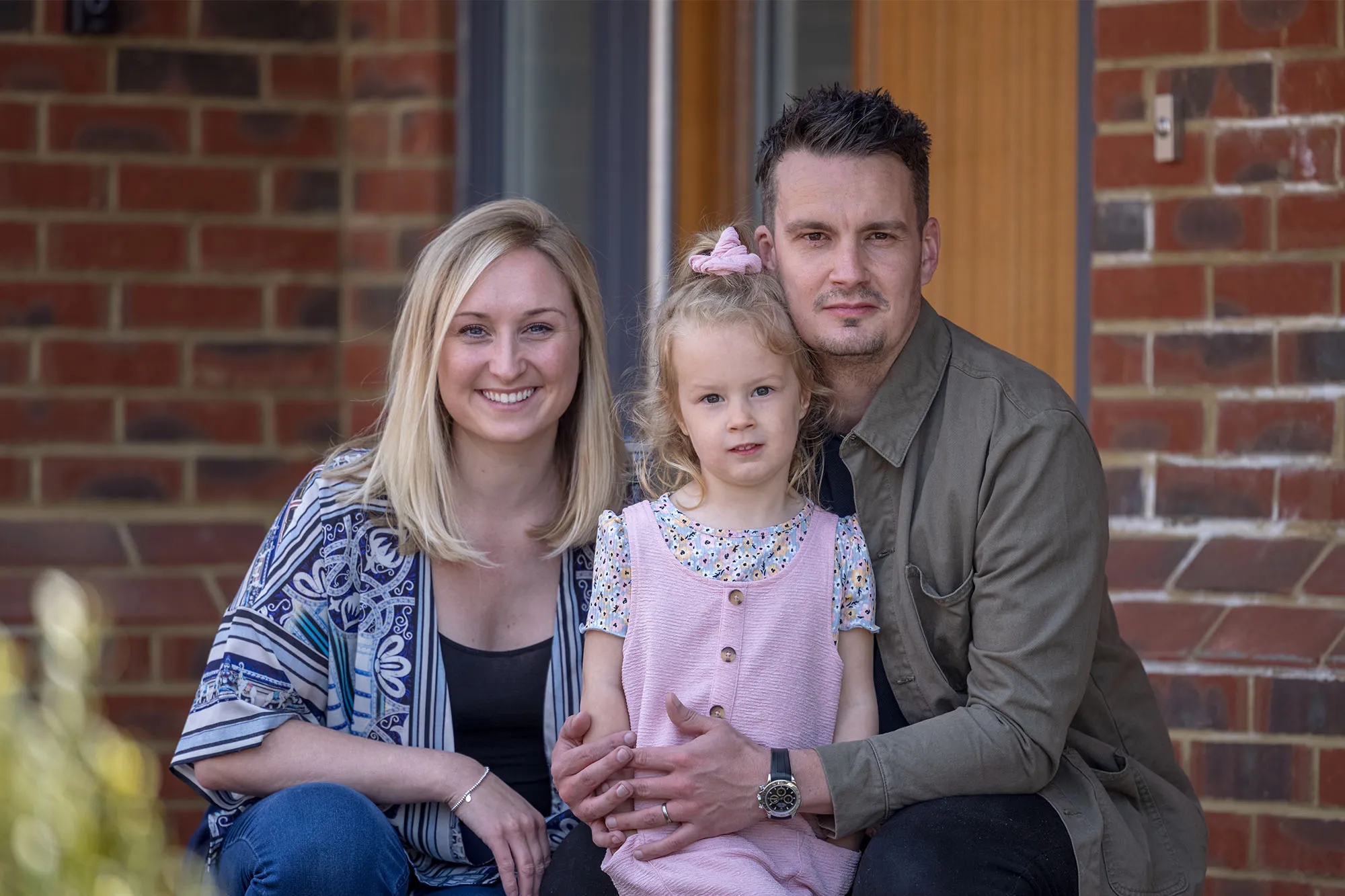 Three people sit closely together on outdoor steps in front of a red brick wall and white door. The person on the left wears a blue-and-white patterned blouse with jeans, the center figure wears a pink dress layered over a floral shirt, and the person on the right wears a green T-shirt with dark trousers and a watch. Their relaxed posture and proximity suggest a familial or friendly bond.