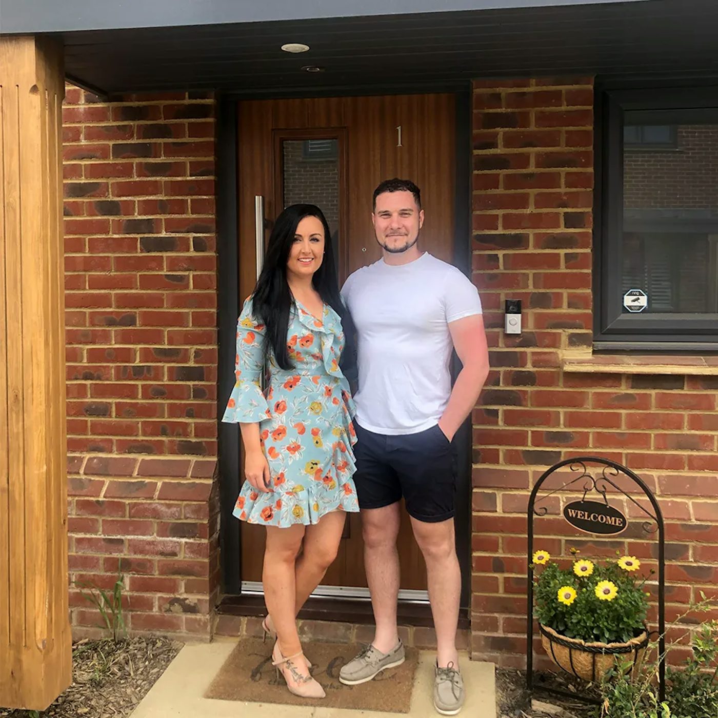 Two people stand near the entrance of a brick house, which features a wooden front door and a window to its right. A small garden with a ‘Welcome’ sign and a pot of yellow flowers adds a cheerful, inviting touch to the scene.
