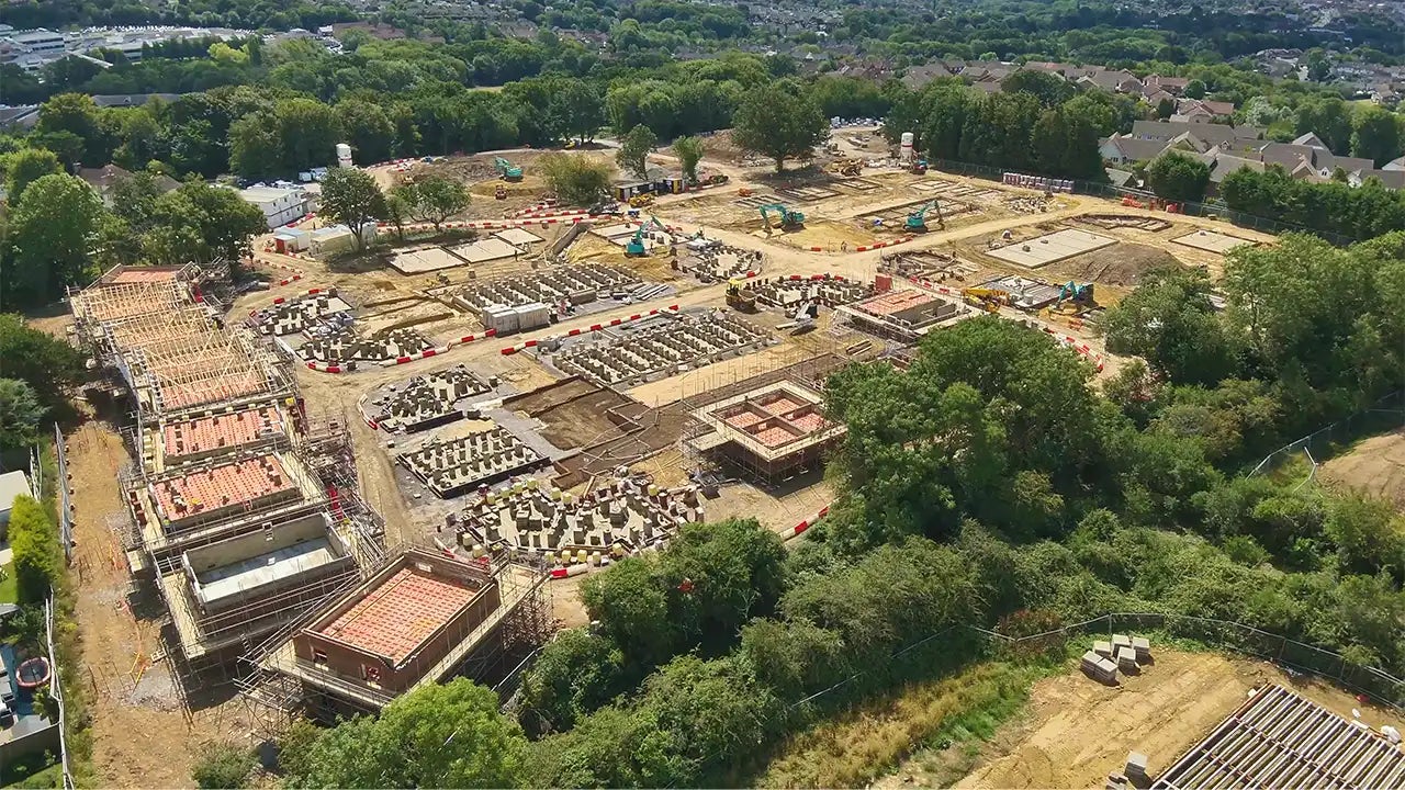 An expansive construction site bordered by mature trees and residential neighborhoods. Several multi-story buildings are in various stages of development, with exposed brickwork, scaffolding, and concrete frames visible. Construction vehicles, equipment, and workers are dispersed across the site, highlighting active progress and the large scale of the project. The surrounding greenery provides a striking contrast to the industrial activity within.