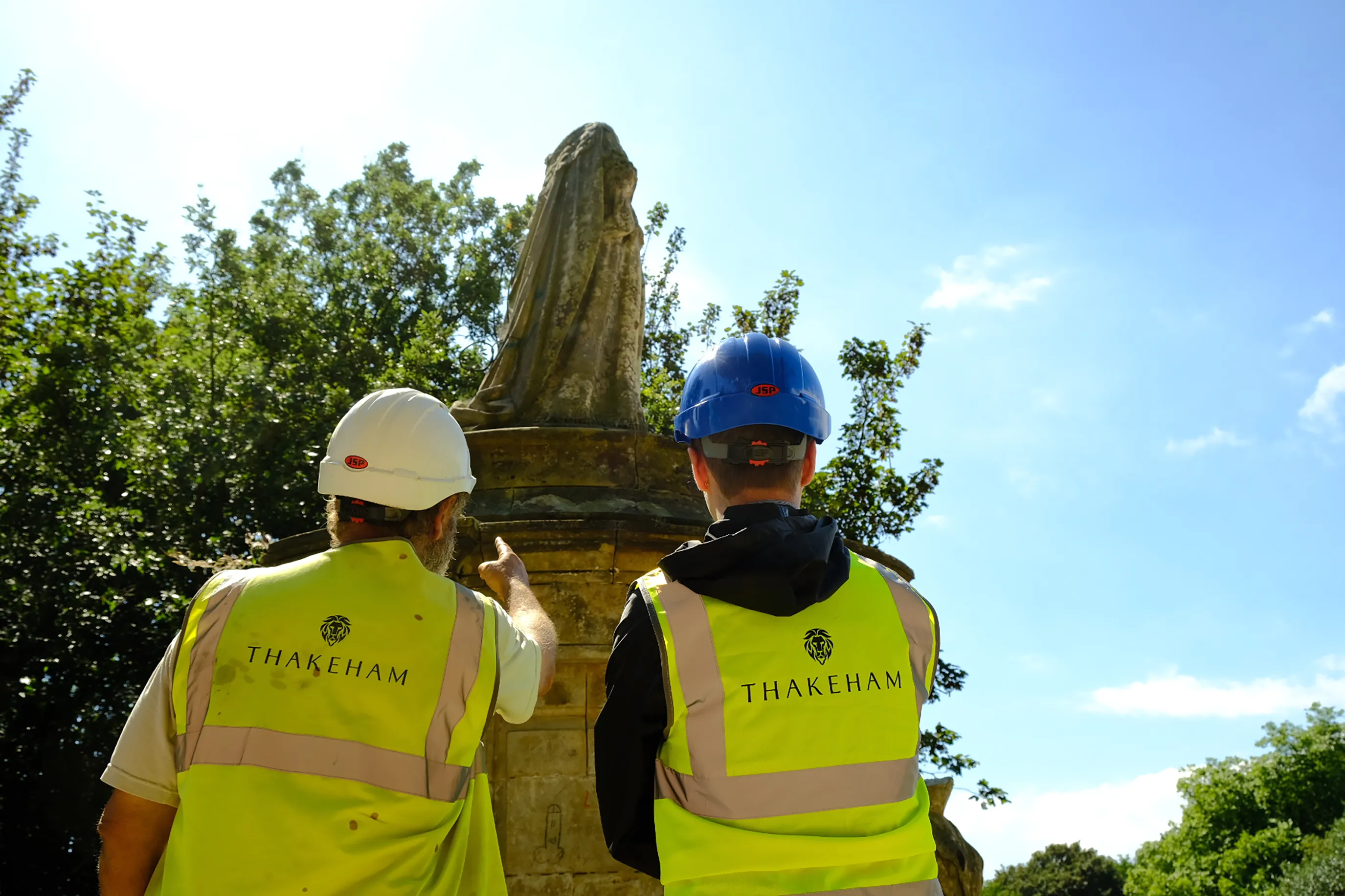 Two individuals in high-visibility vests and white hard hats stand in front of a stone monument or statue, with one person gesturing toward it. The vests are branded with "THAKEHAM" on the back, suggesting involvement in construction or site evaluation. Trees and a clear blue sky provide a calm, natural backdrop.