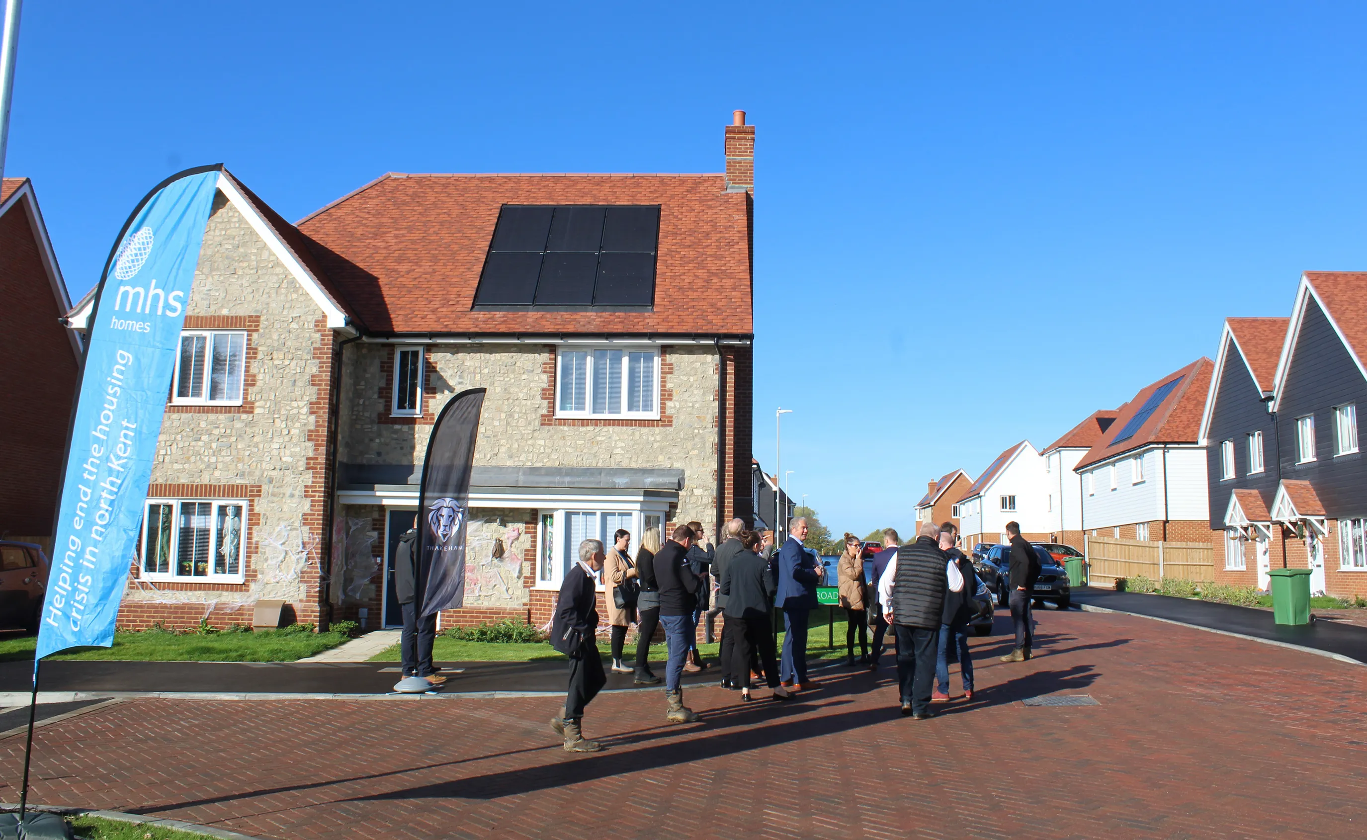 A group of people gathered outside a modern house with rooftop solar panels, participating in a daytime community event. Two branded flags are visible—one displaying 'mhs homes' and the other with the phrase 'Helping on the housing crisis in north Kent.' Similar homes line the residential street in the background, suggesting a focus on housing solutions and sustainability.